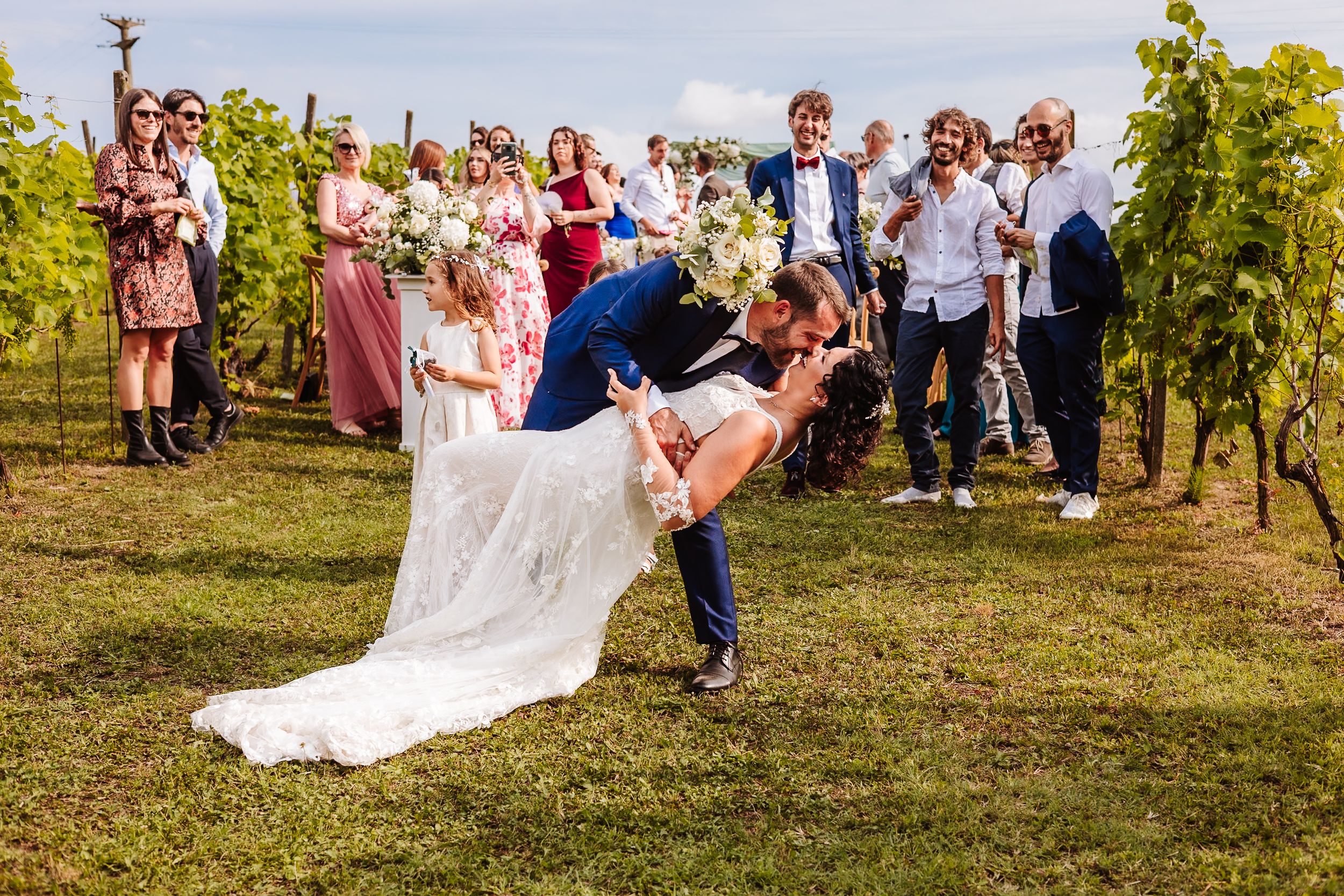 Groom dips bride for a kiss at outdoor vineyard wedding while guests cheer and flower girl watches nearby.