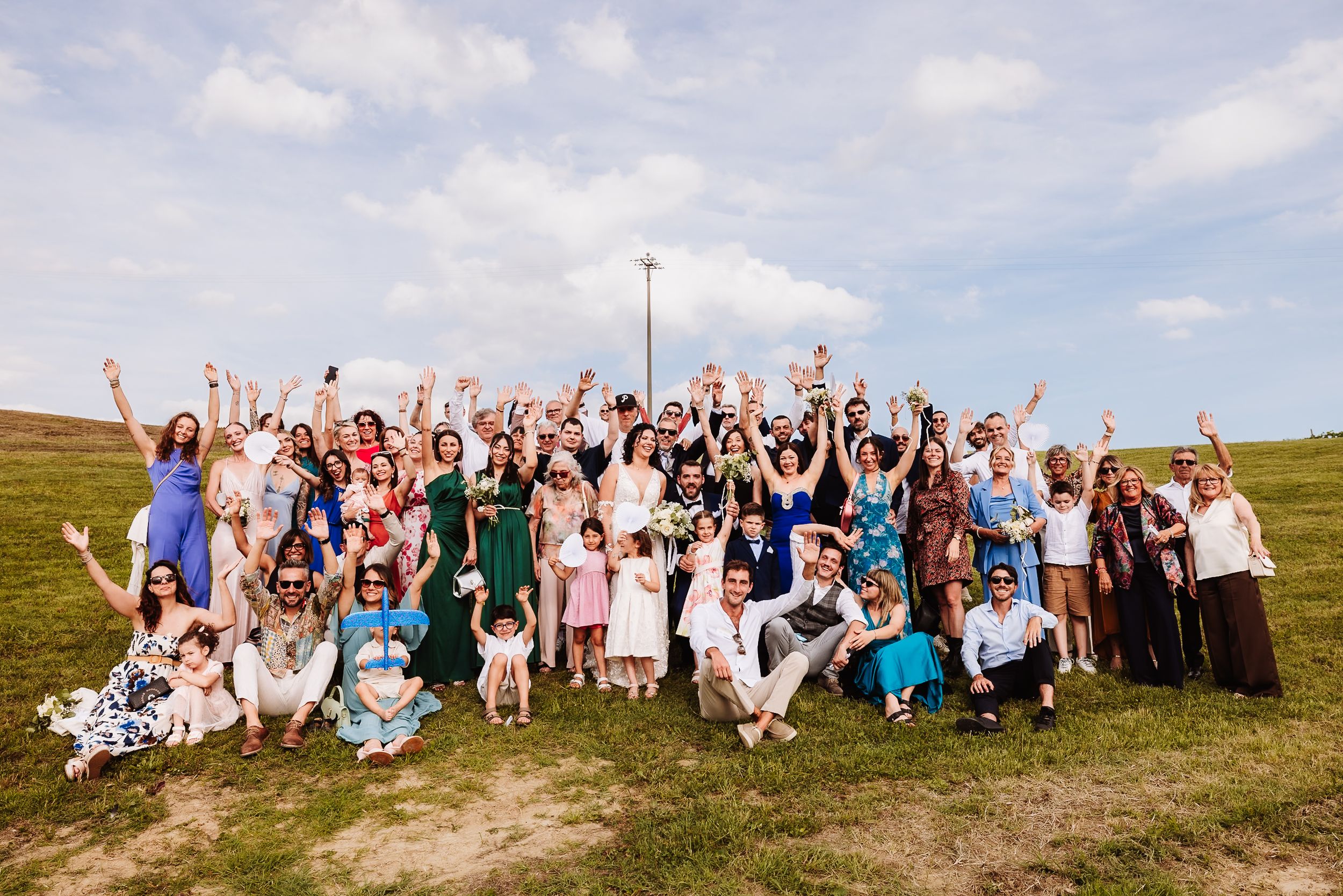 Large wedding group photo outdoors with guests cheering and raising hands on a green hillside under a blue sky.