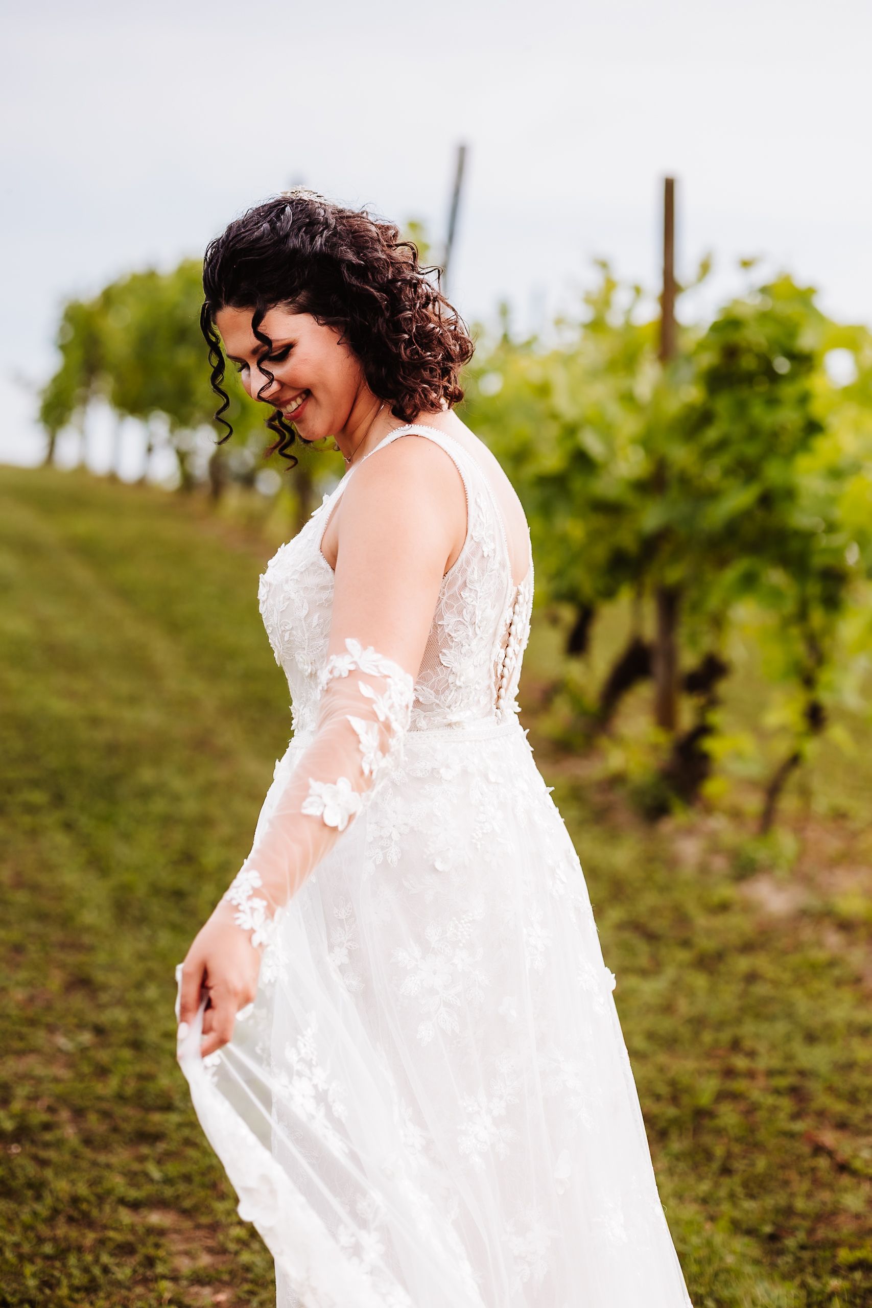 Bride in lace wedding gown smiling in vineyard.