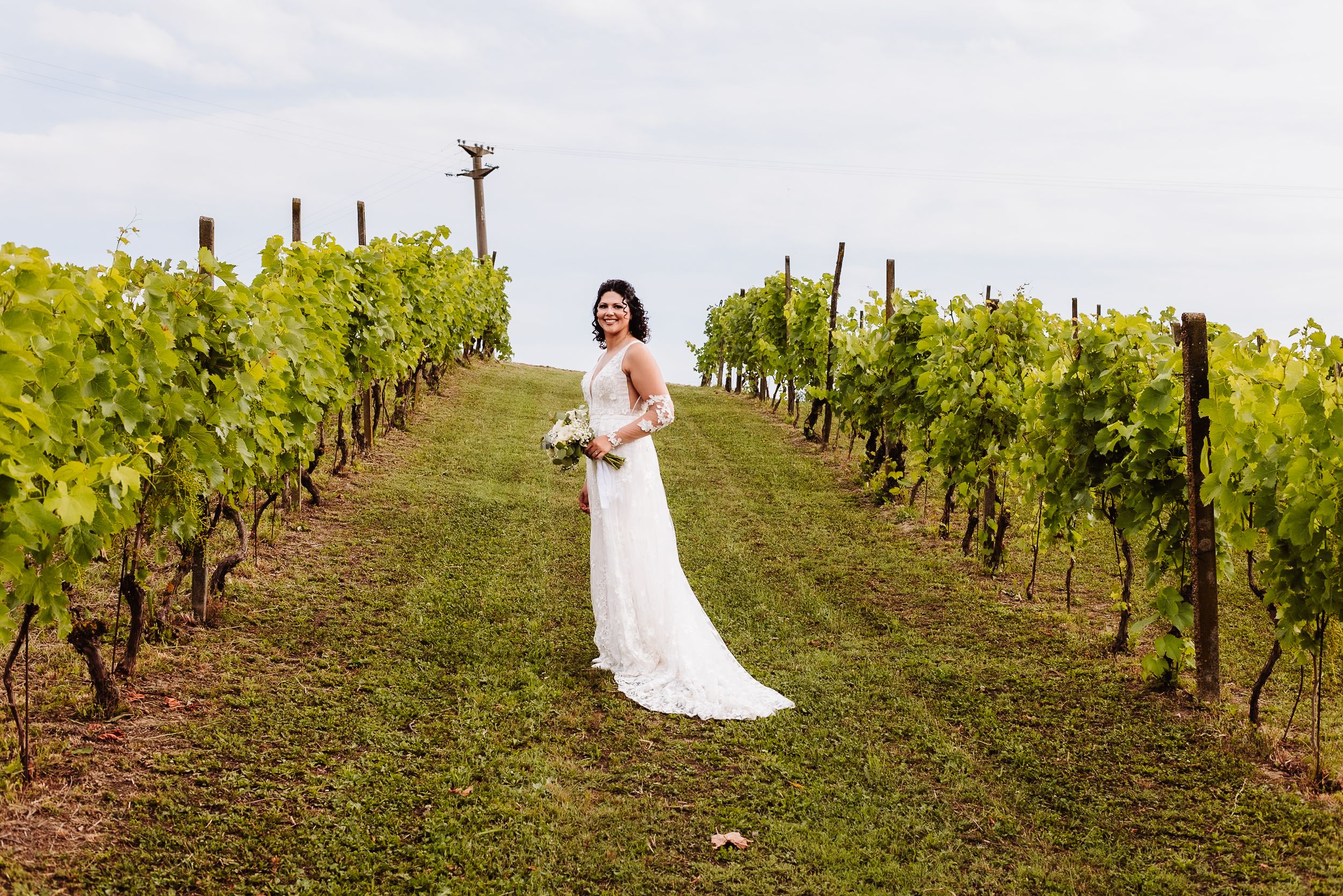 Bride in white gown standing in lush vineyard rows, holding bouquet, smiling on her wedding day.