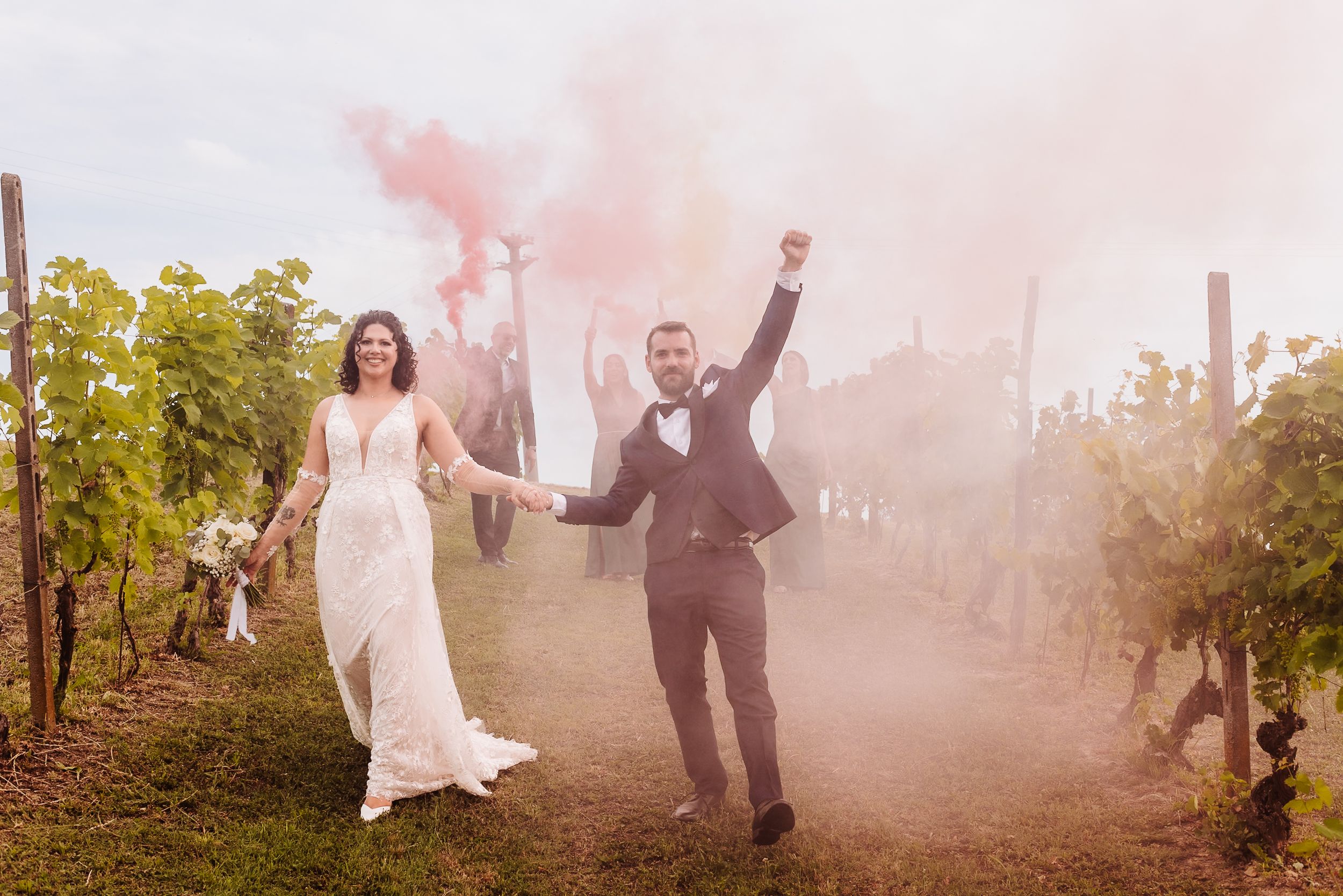 Bride and groom celebrate with pink smoke bomb in vineyard, groom raising arm joyfully on wedding day.