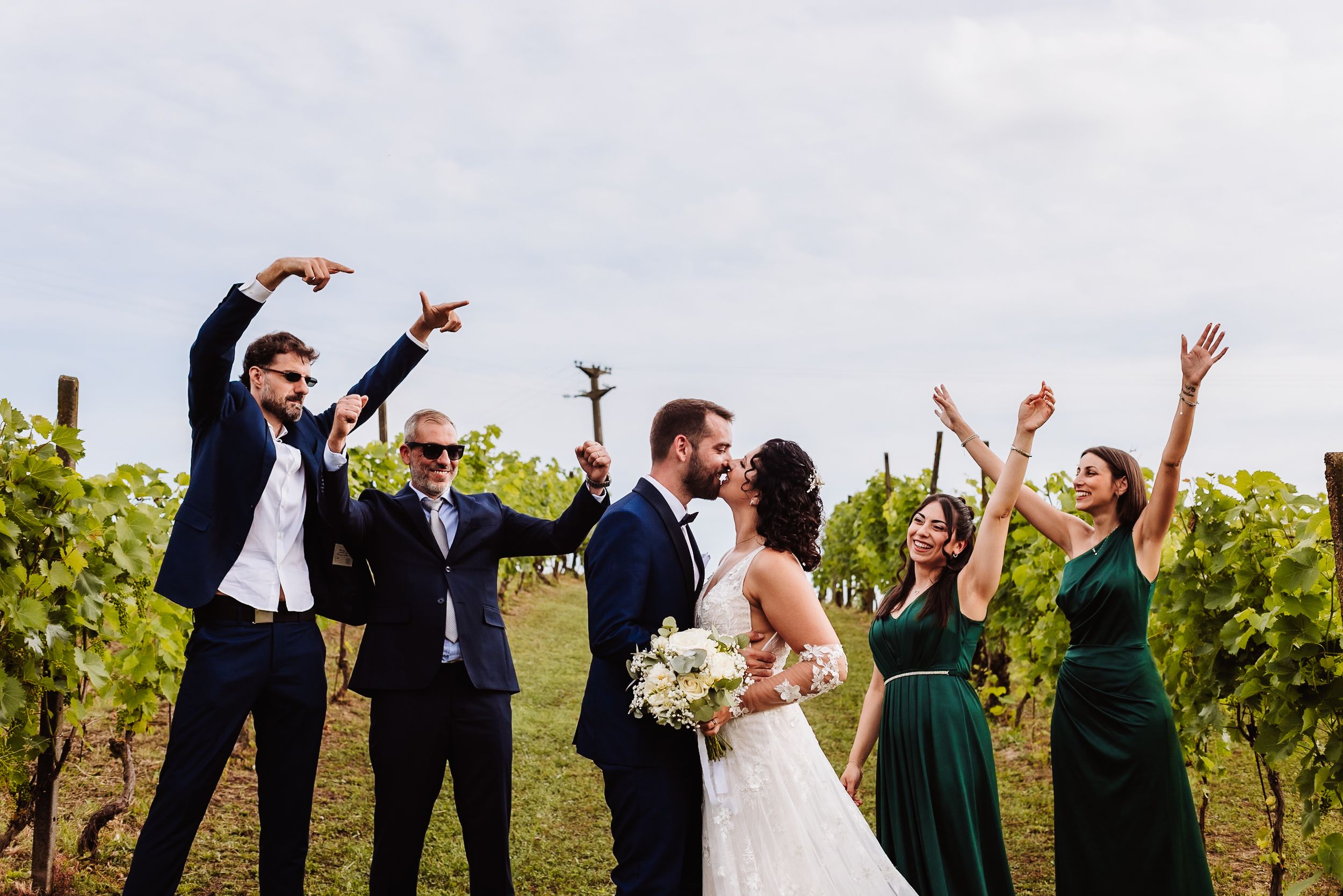 Wedding party celebrating in a vineyard, bride and groom kissing while guests cheer in formal attire.