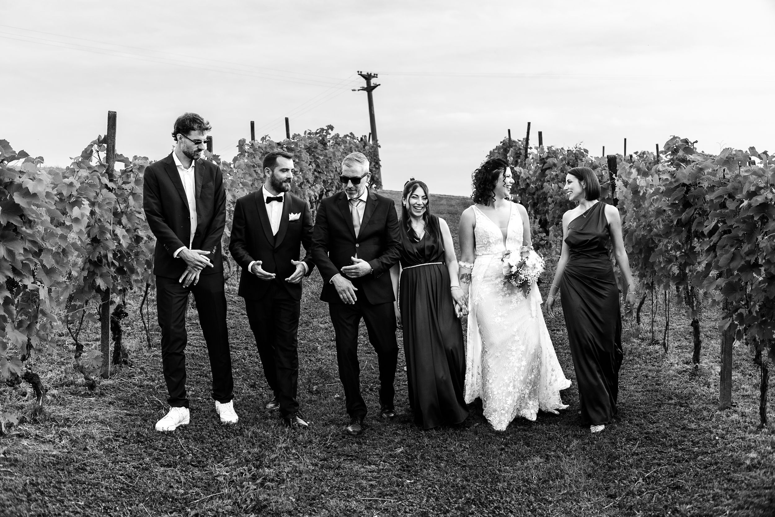 Black and white photo of wedding party walking through vineyard rows, bride in lace gown with bouquet.