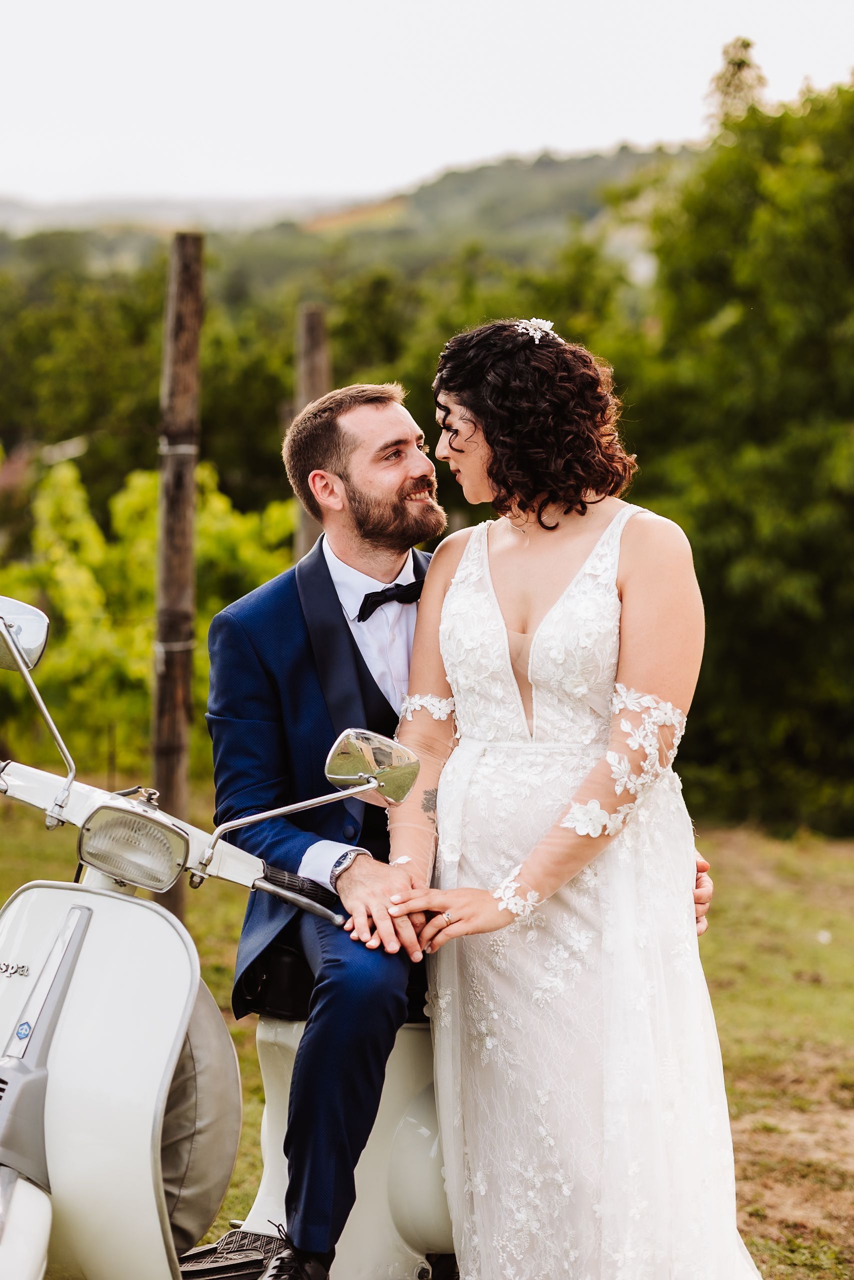 A bride and groom share a romantic moment beside a white Vespa scooter amid lush green vineyard scenery.