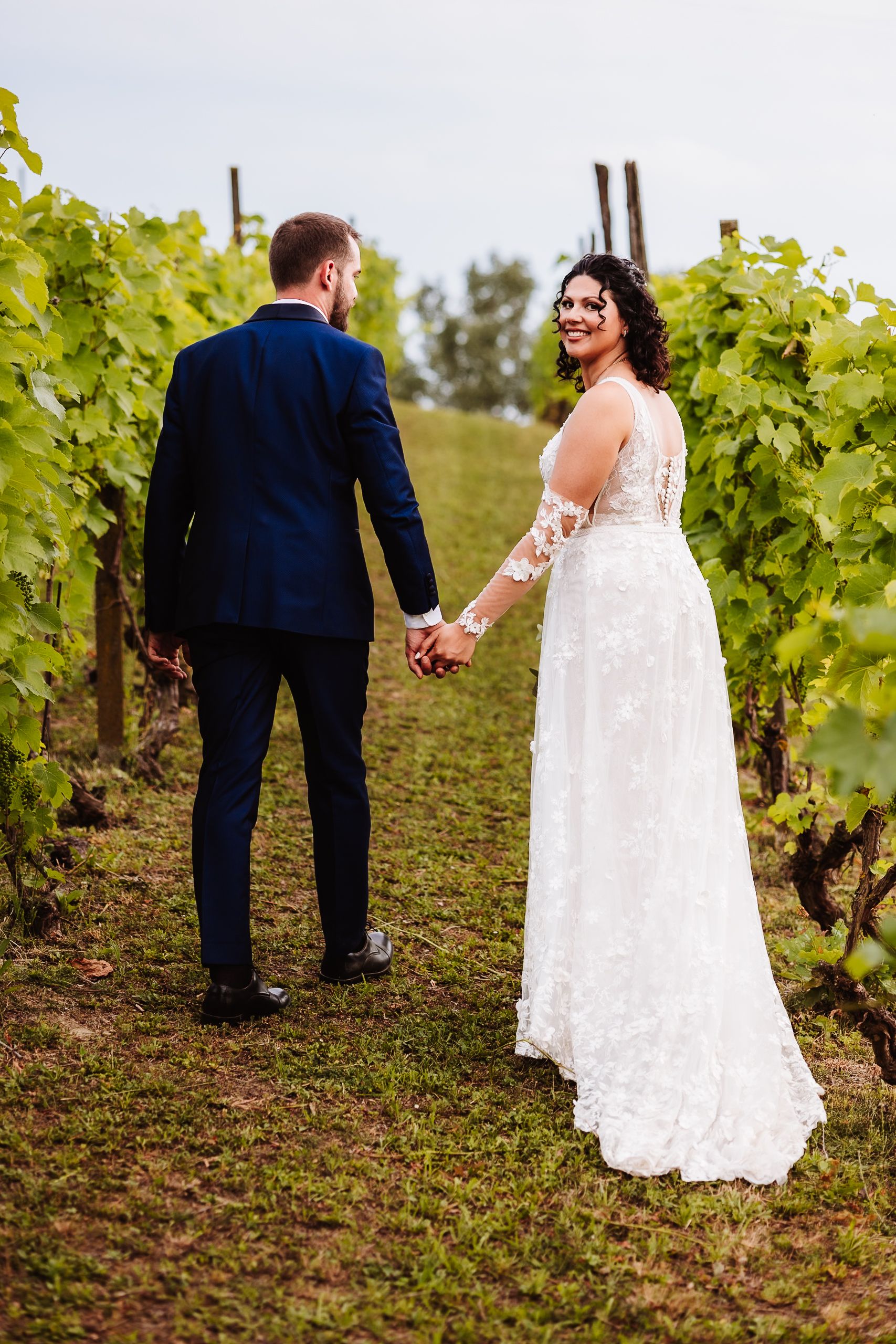 Bride and groom walking hand in hand through a lush vineyard, bride smiling back in a lace gown.