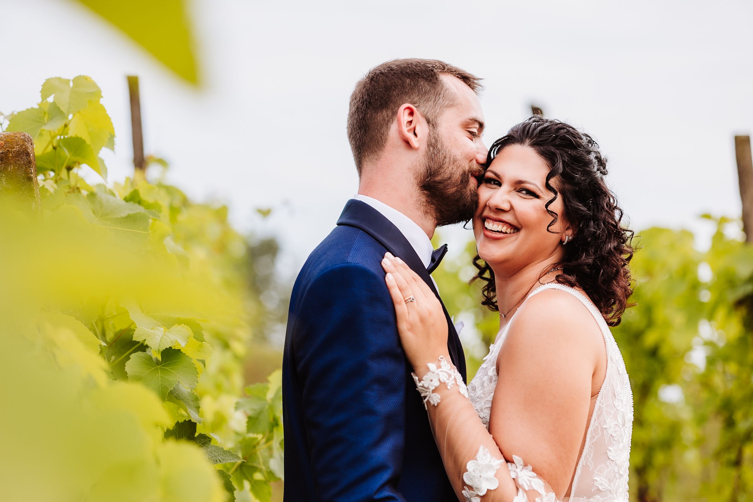 A bride and groom share a romantic moment among lush vineyard vines, smiling and embracing on their wedding day.