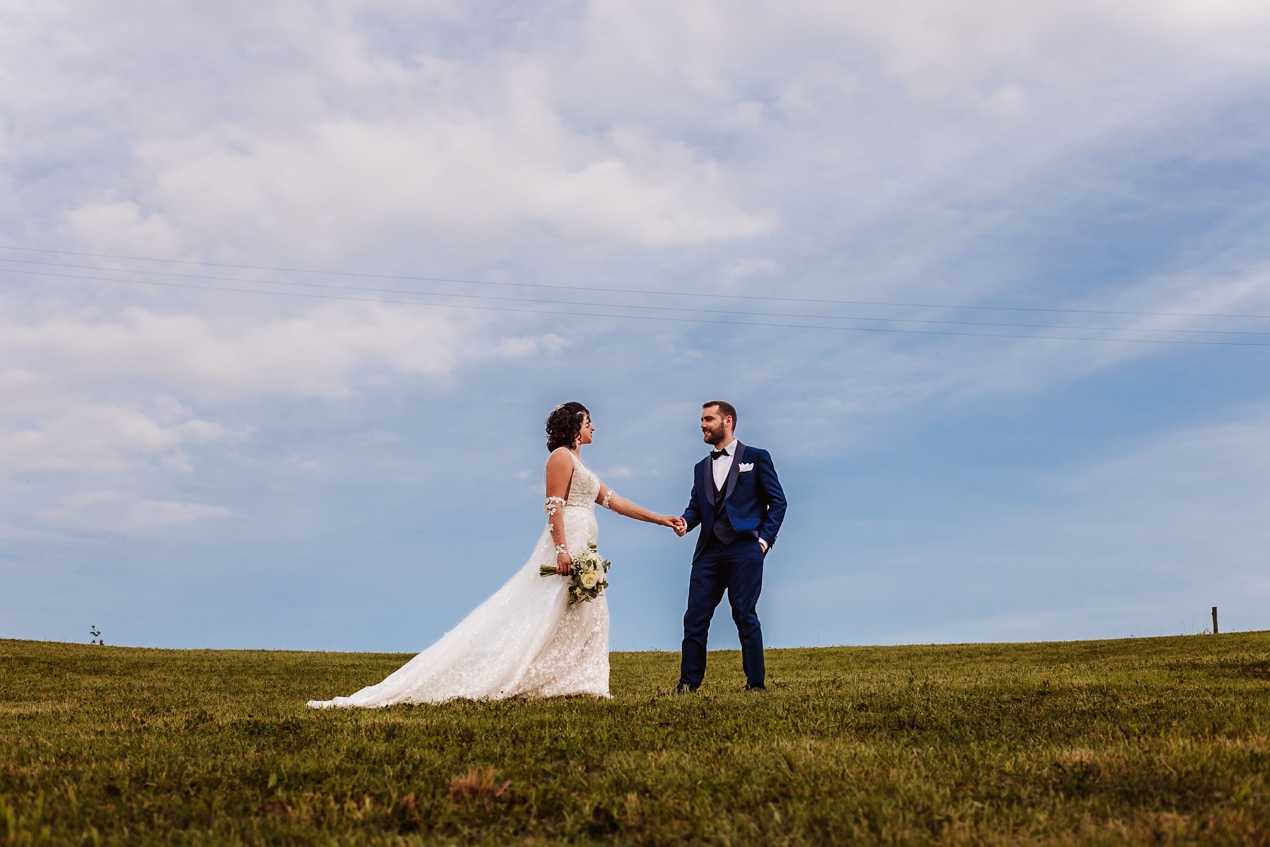 Bride and groom holding hands on a green hillside under a blue sky on their wedding day.
