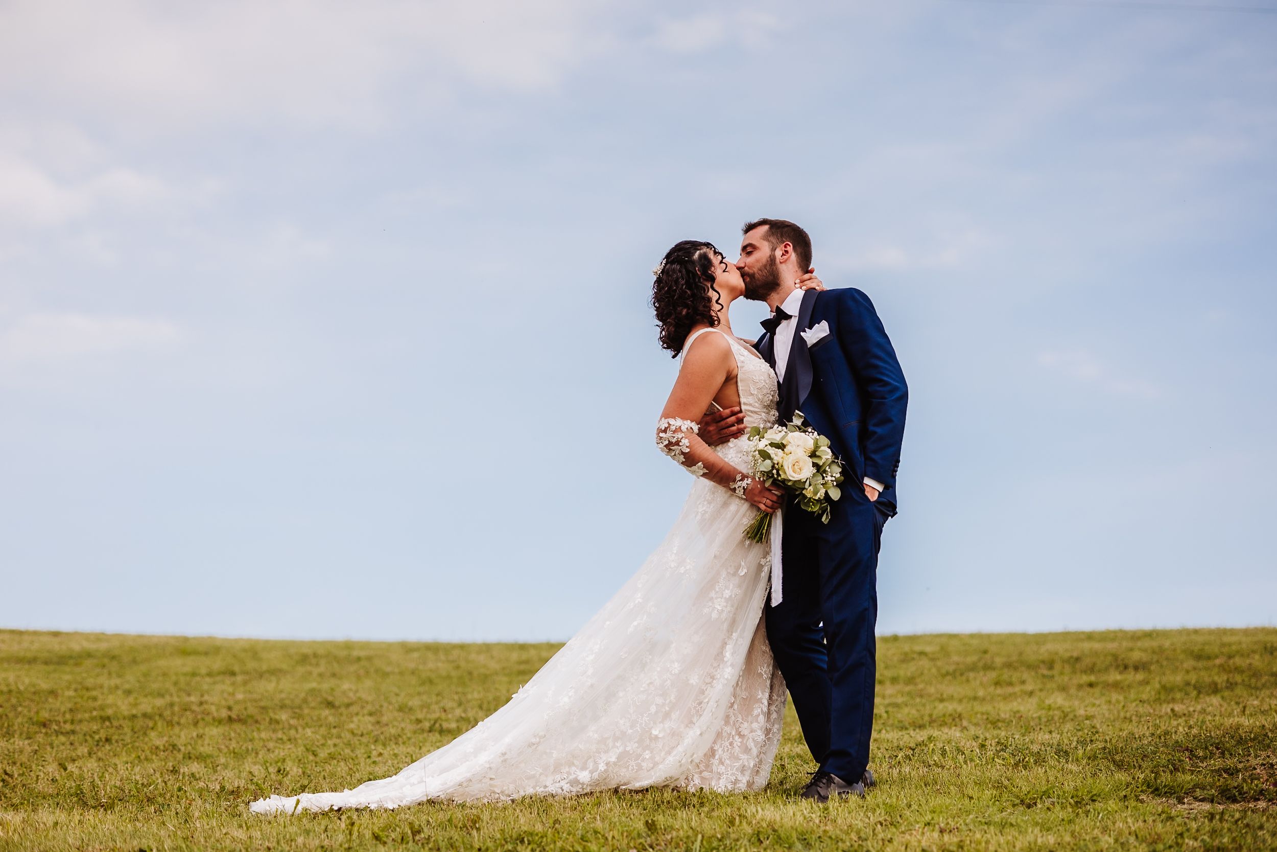Bride and groom share a romantic kiss on a green lawn under a blue sky on their wedding day.