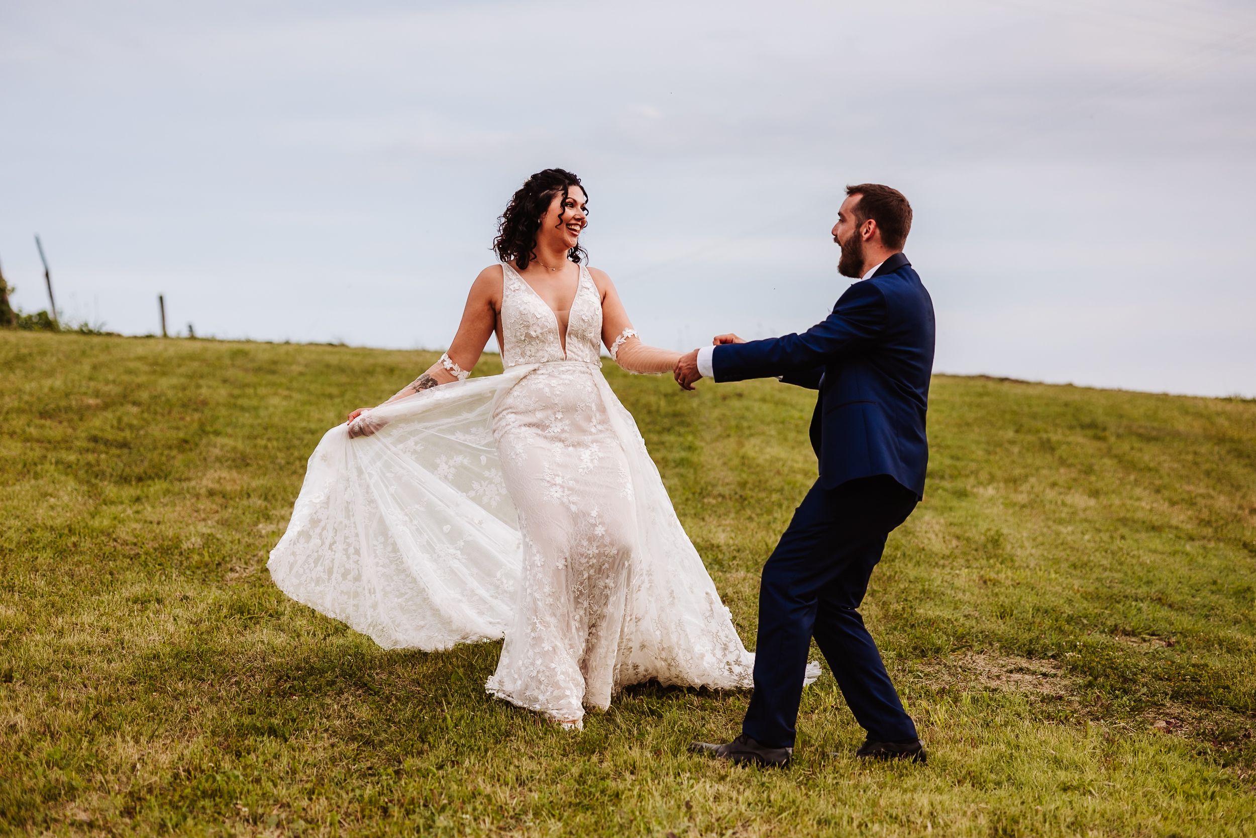 A bride in a lace gown twirls with her groom in a navy suit on a green hillside during their wedding photos.