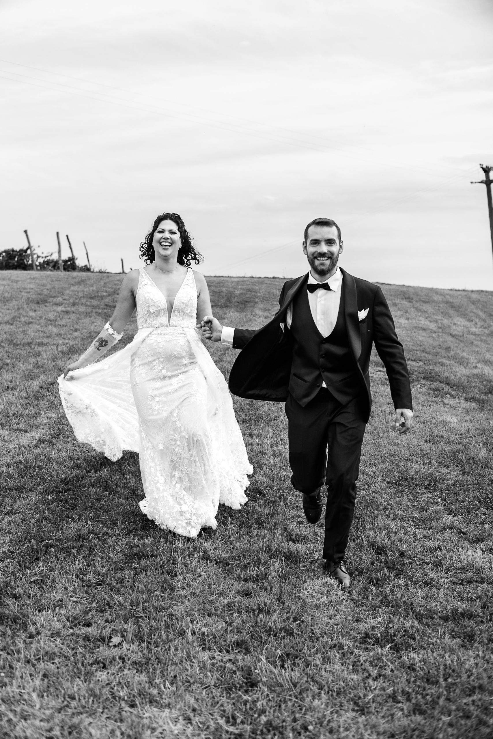 Black and white photo of a laughing bride and groom holding hands running across a grassy field on their wedding day.