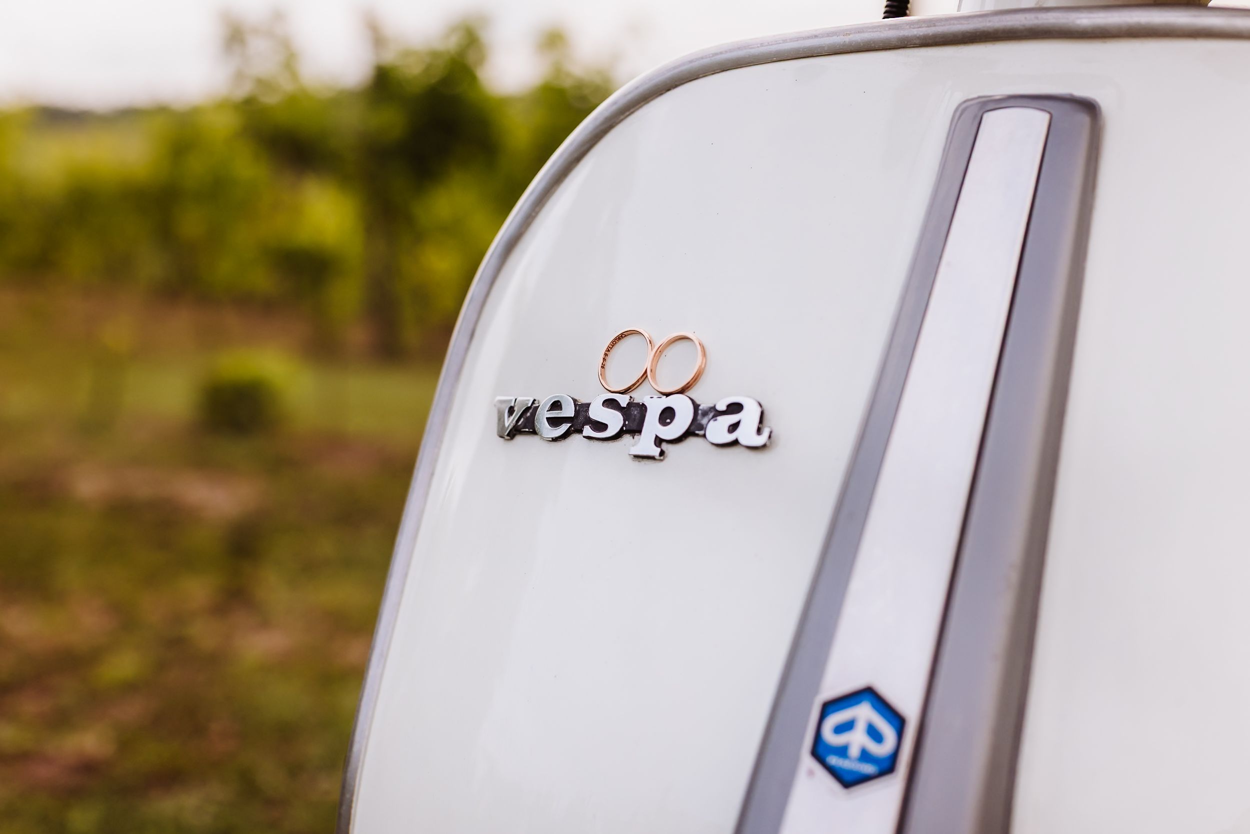 Close-up of a white Vespa scooter badge logo with green vineyard background.