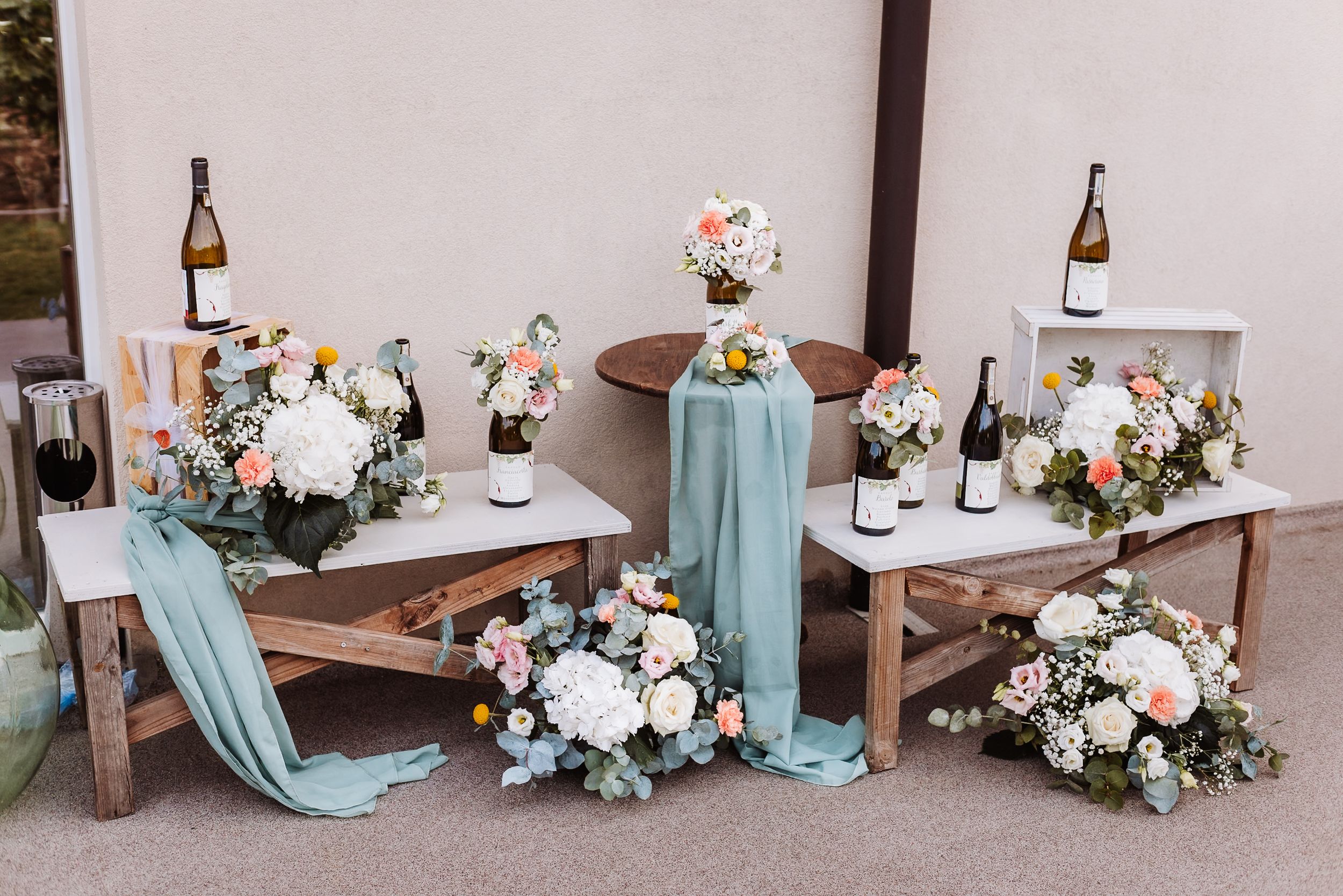 Elegant wedding sweetheart table with teal fabric, white floral arrangements, and wine bottles on rustic wooden benches.