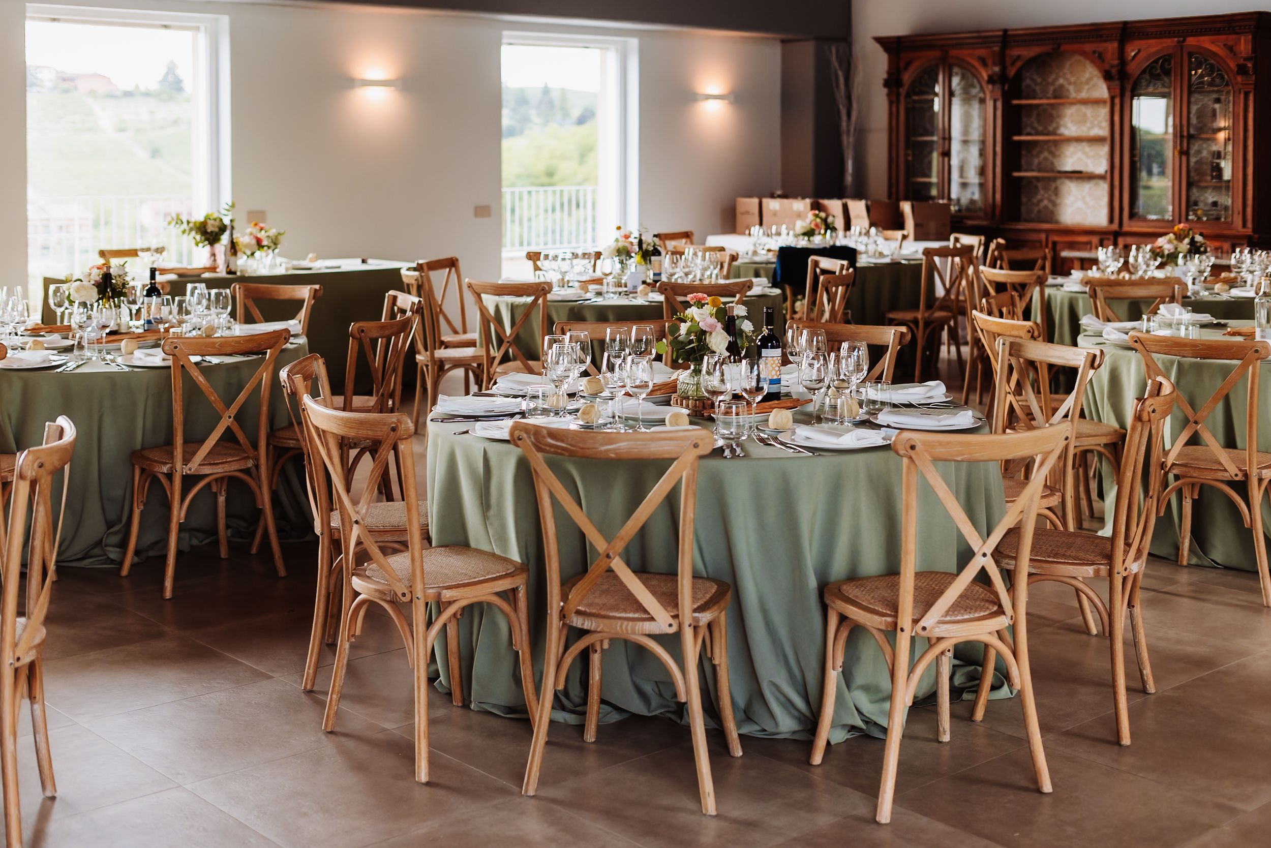 Elegant wedding reception hall with sage green tablecloths, wooden cross-back chairs, and floral centerpieces on round tables.