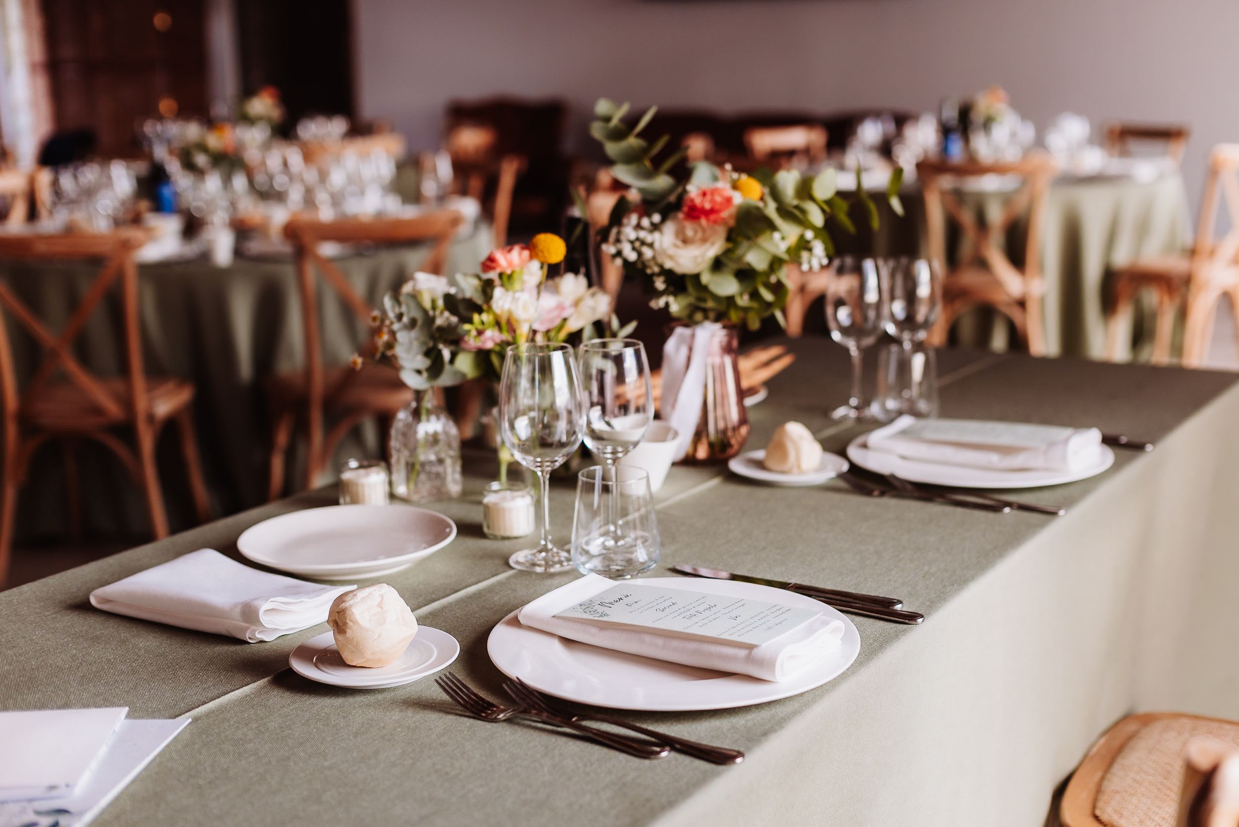 Elegant wedding reception table setting with sage green linen, white plates, glassware, and colorful floral centerpieces.