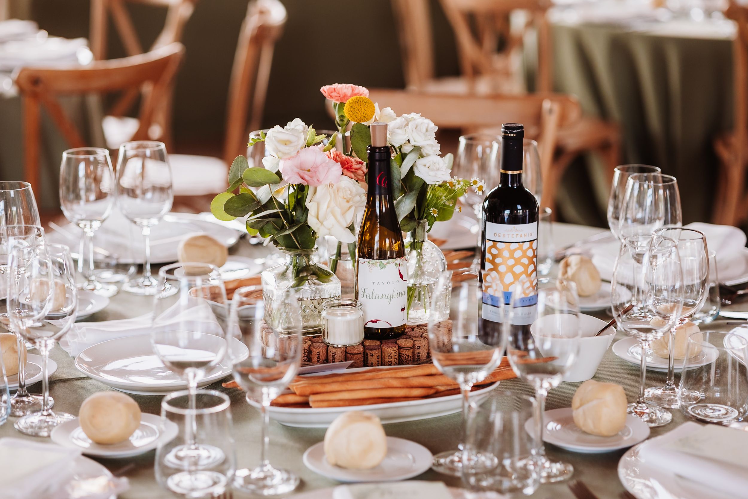 Elegant wedding reception table with floral centerpiece, wine bottles, and crystal glassware on white linen.