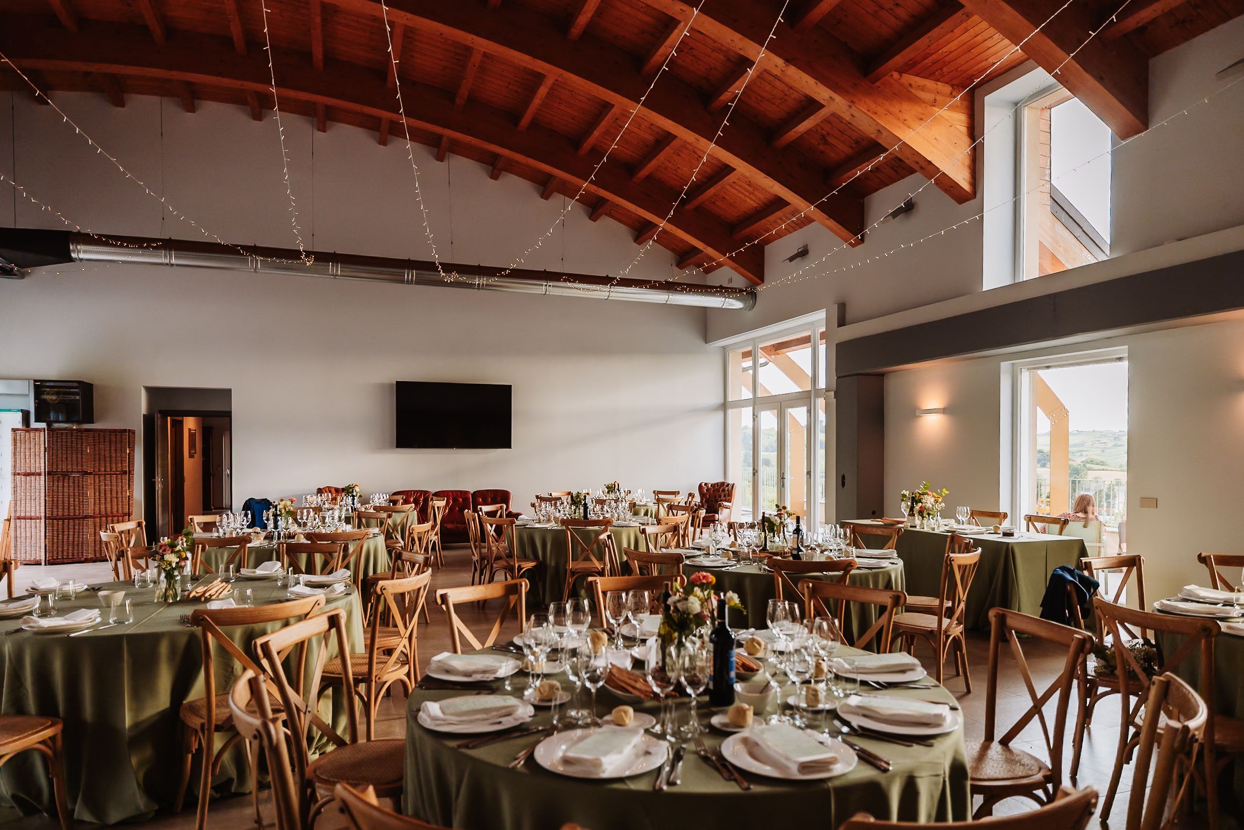 Elegant wedding reception hall with wooden vaulted ceiling, round tables with sage green linens, and cross-back chairs.