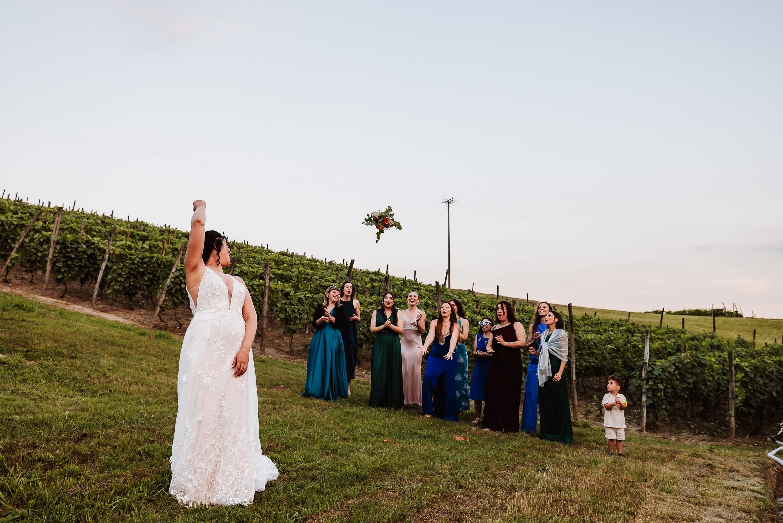 Bride tossing bouquet to bridesmaids in colorful gowns at outdoor vineyard wedding ceremony.