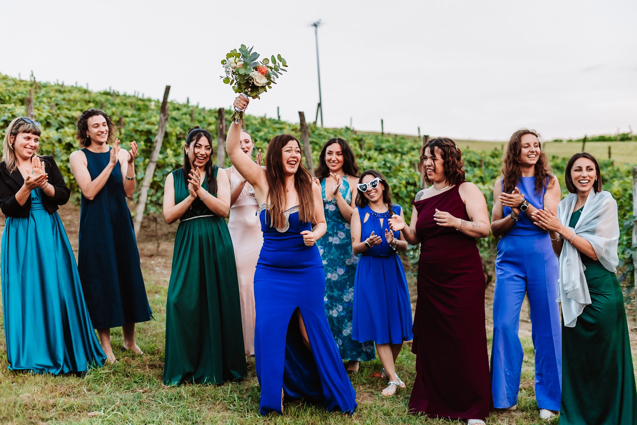 Bridesmaids in colorful blue, teal, and burgundy gowns celebrate outdoors at a vineyard wedding, clapping as bouquet is tossed.