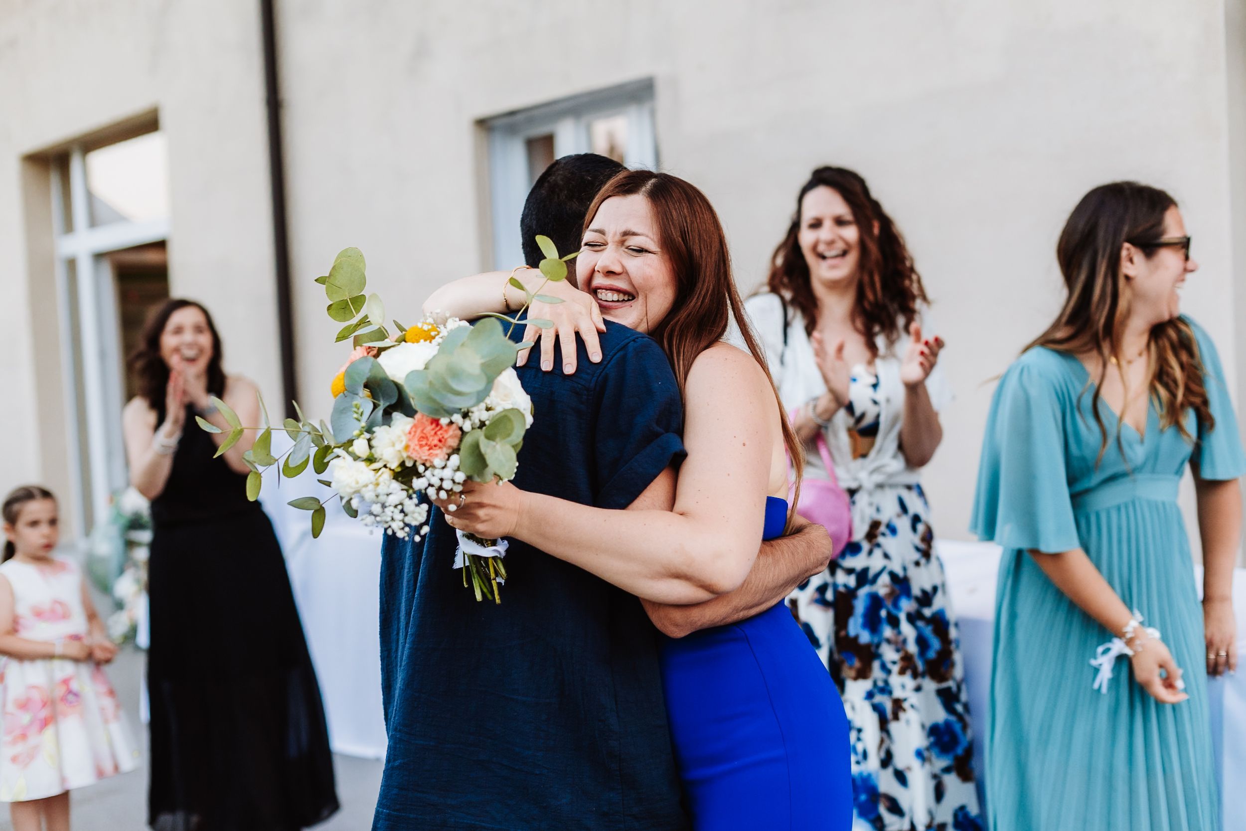 Two women share a joyful hug at a wedding, one holding a bouquet, as guests applaud in the background.