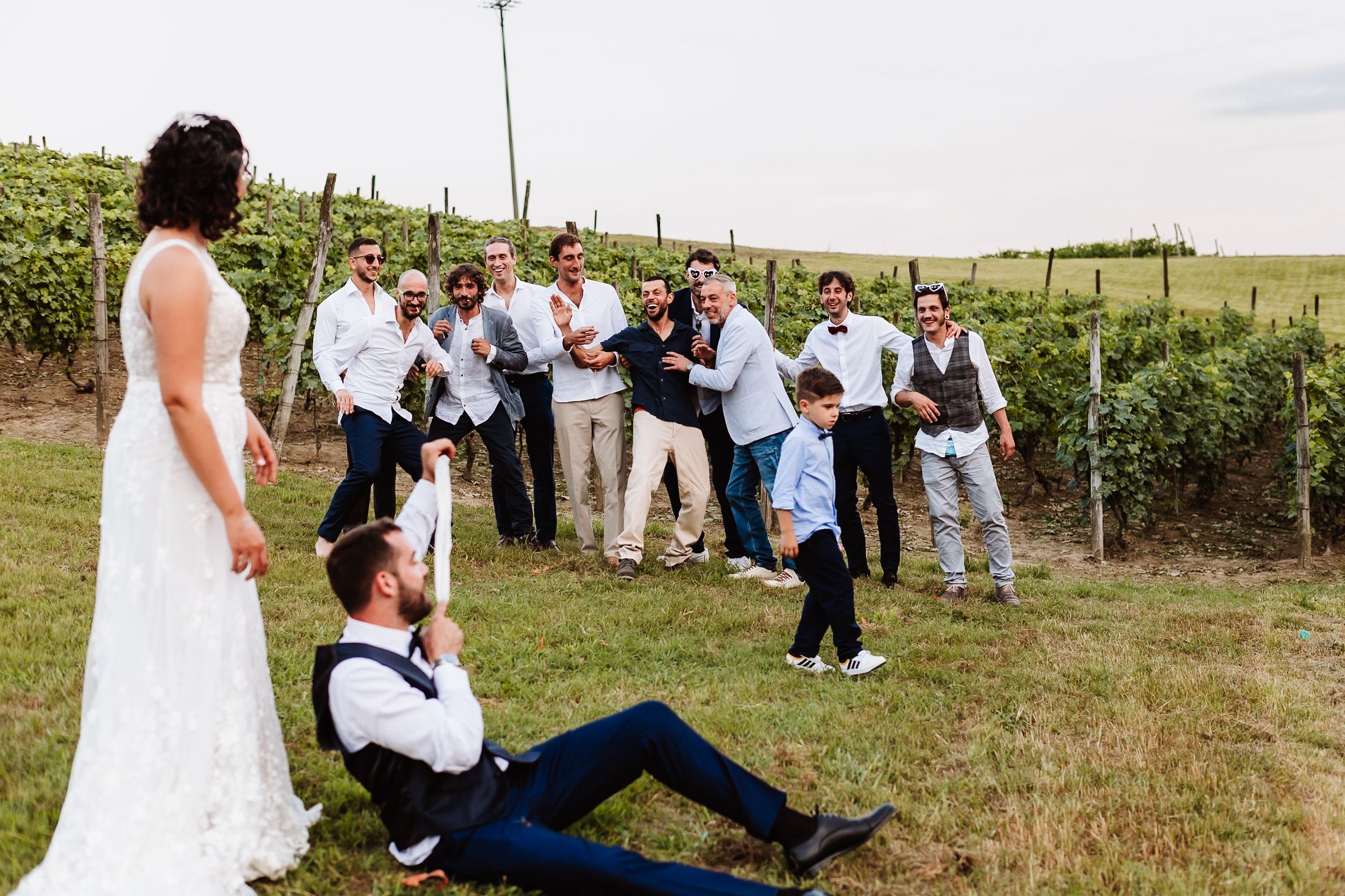 Bride in white gown poses with groom and groomsmen celebrating outdoors at a vineyard wedding.