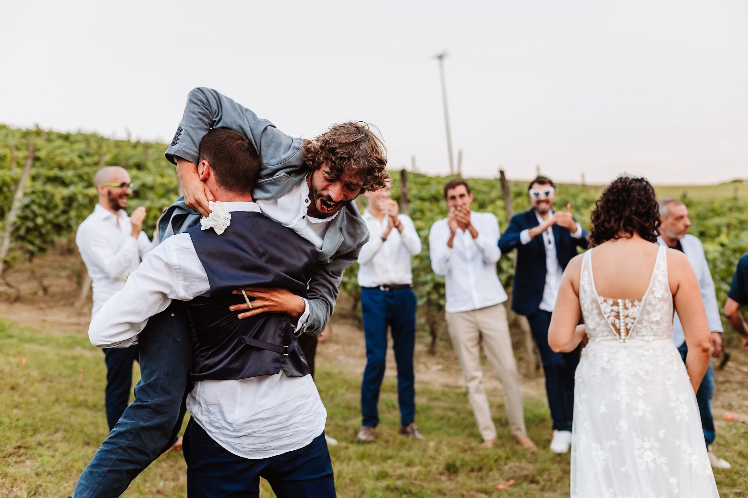 Groom lifted by groomsman at outdoor vineyard wedding celebration with guests applauding in background.