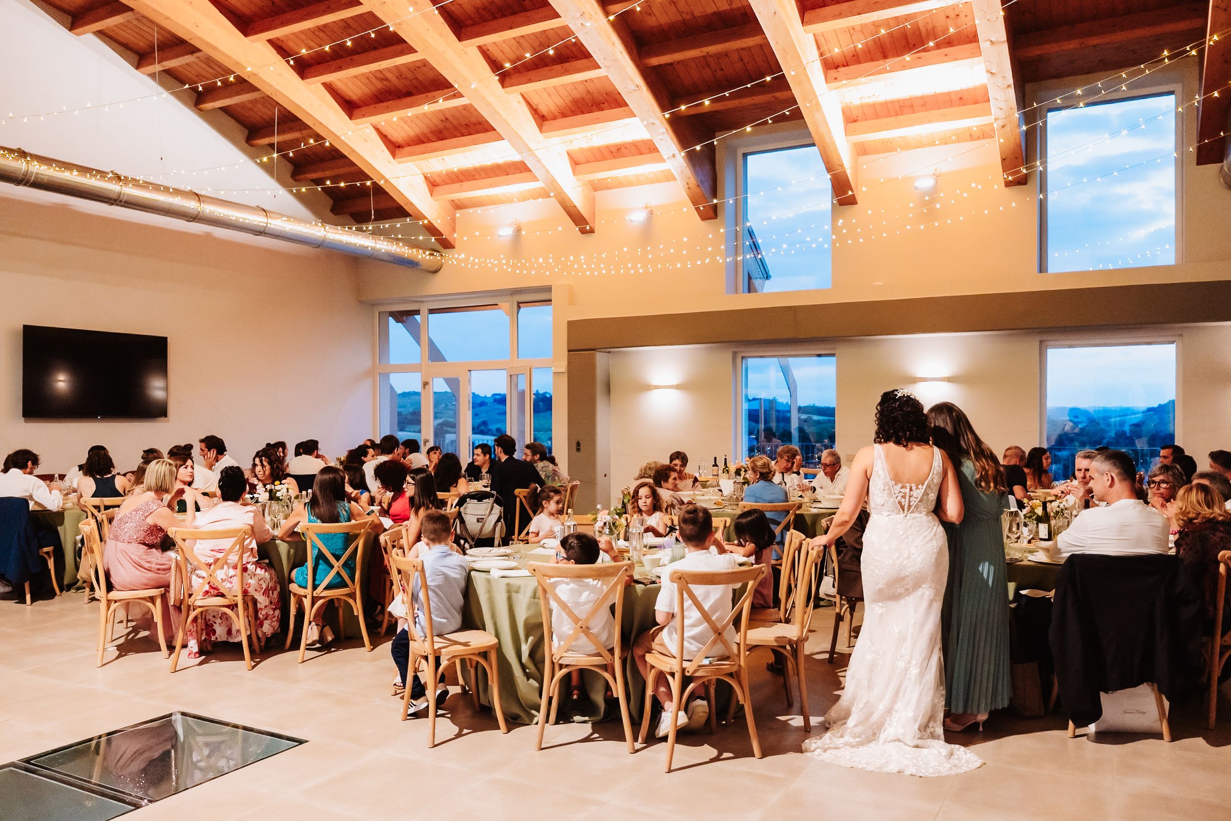Elegant wedding reception in a modern venue with wooden ceiling, fairy lights, and guests seated at round tables.