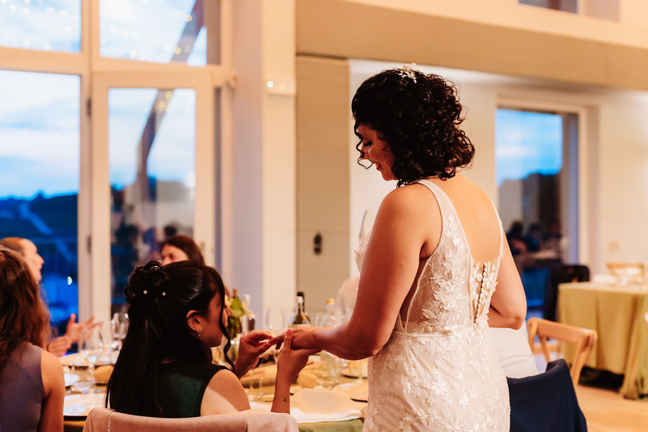 Bride in lace wedding gown interacting with guests at elegant indoor wedding reception with blue evening light.