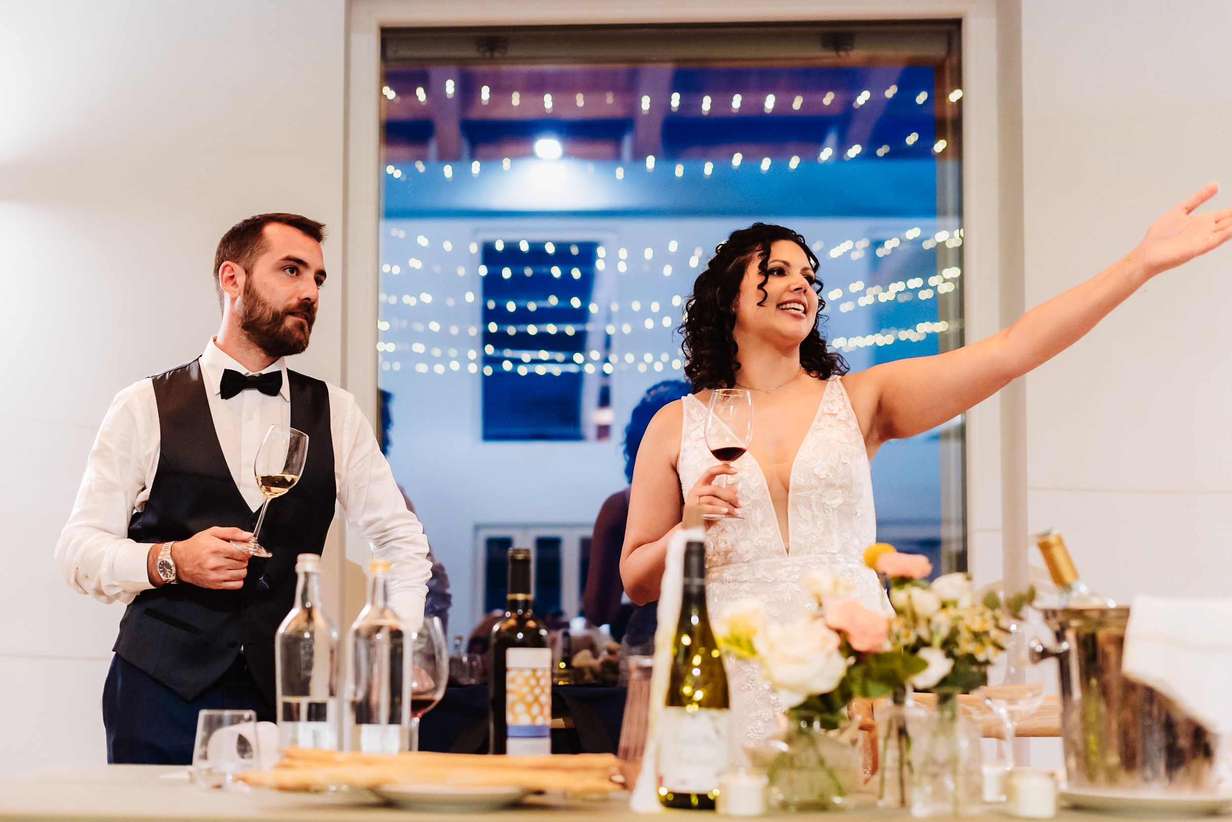 Bride and groom holding wine glasses during wedding reception toast with string lights in background.