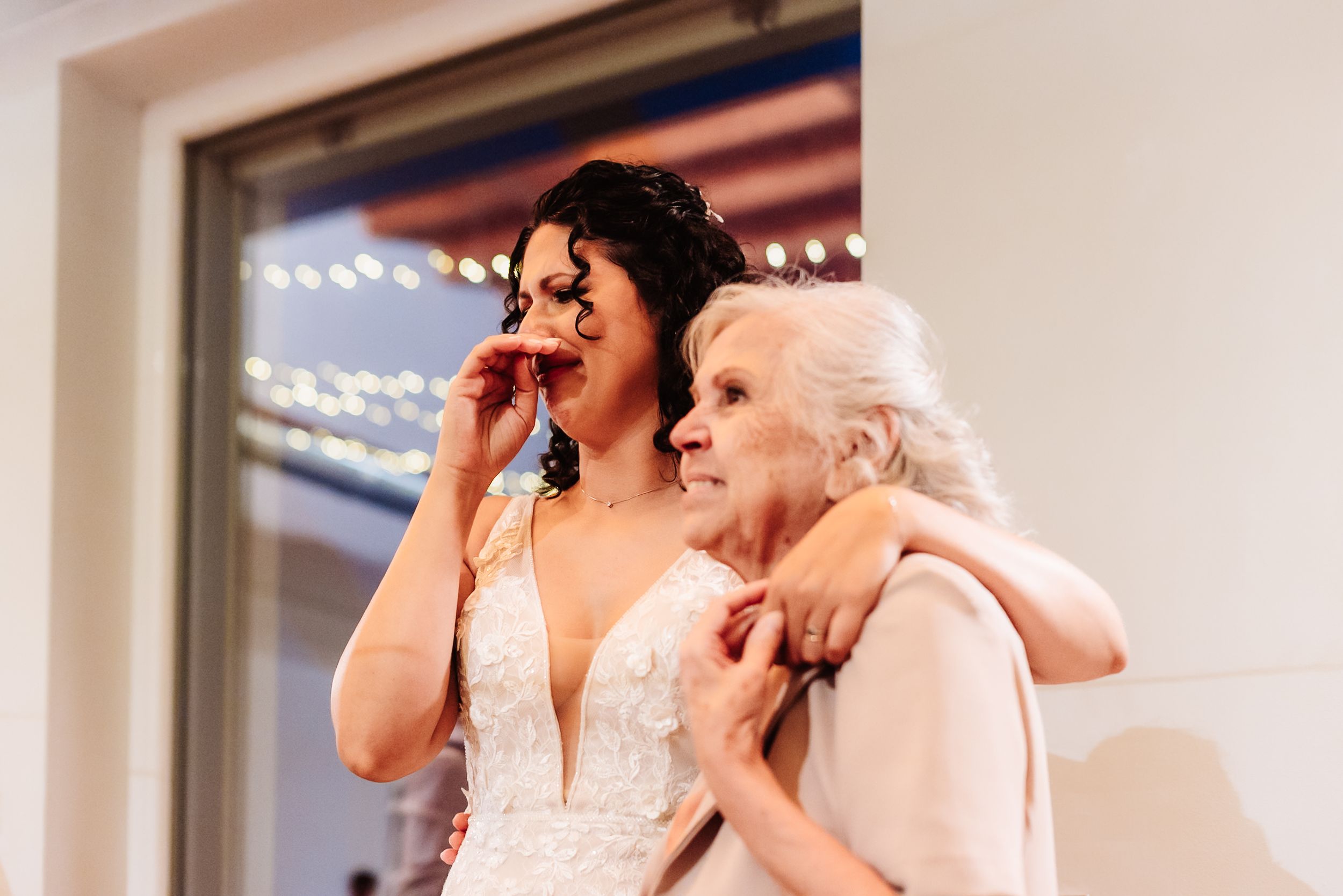 Emotional bride in lace wedding gown wiping tears while embracing elderly woman near window with string lights.