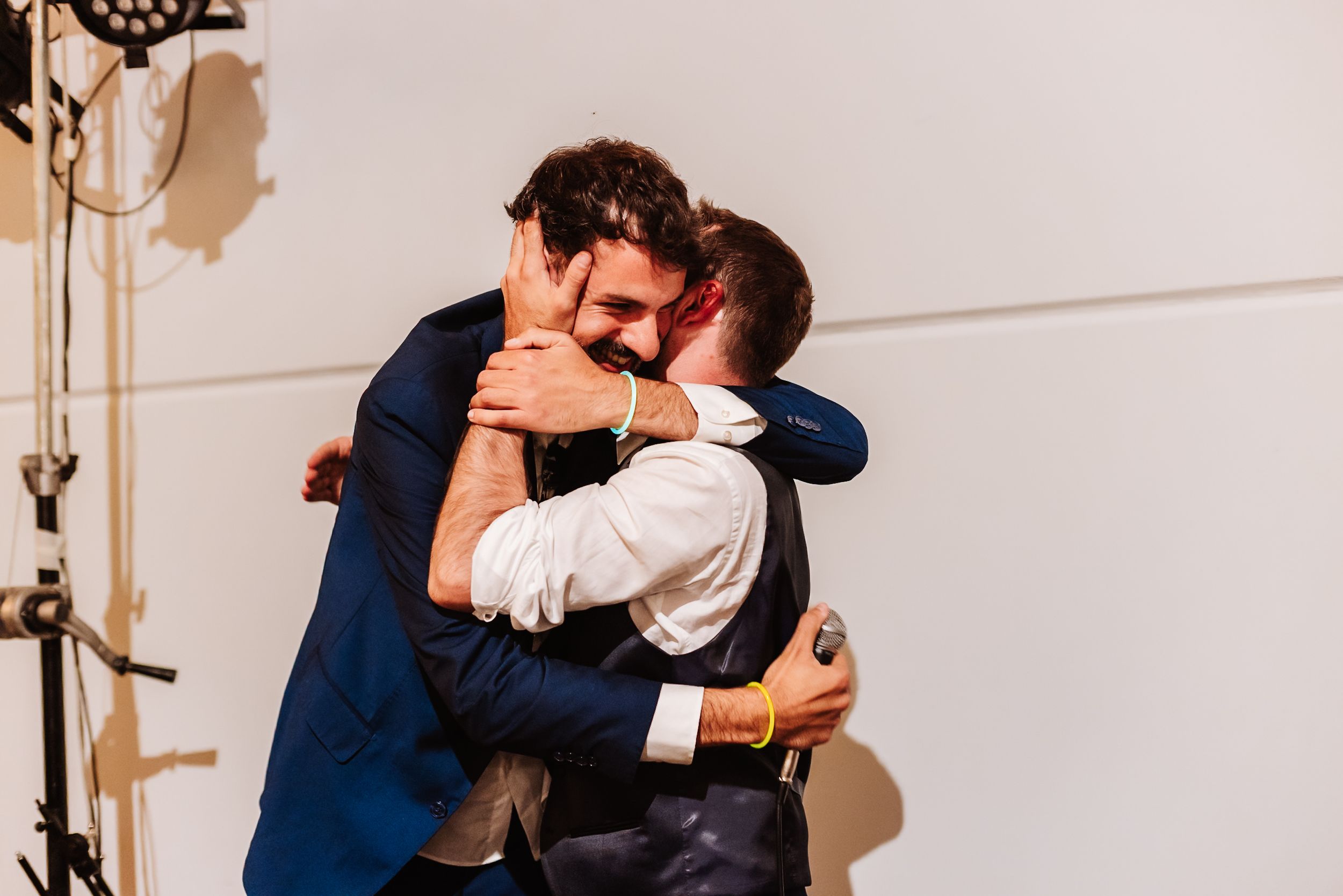Two men in formal attire share a warm embrace at what appears to be a wedding celebration.
