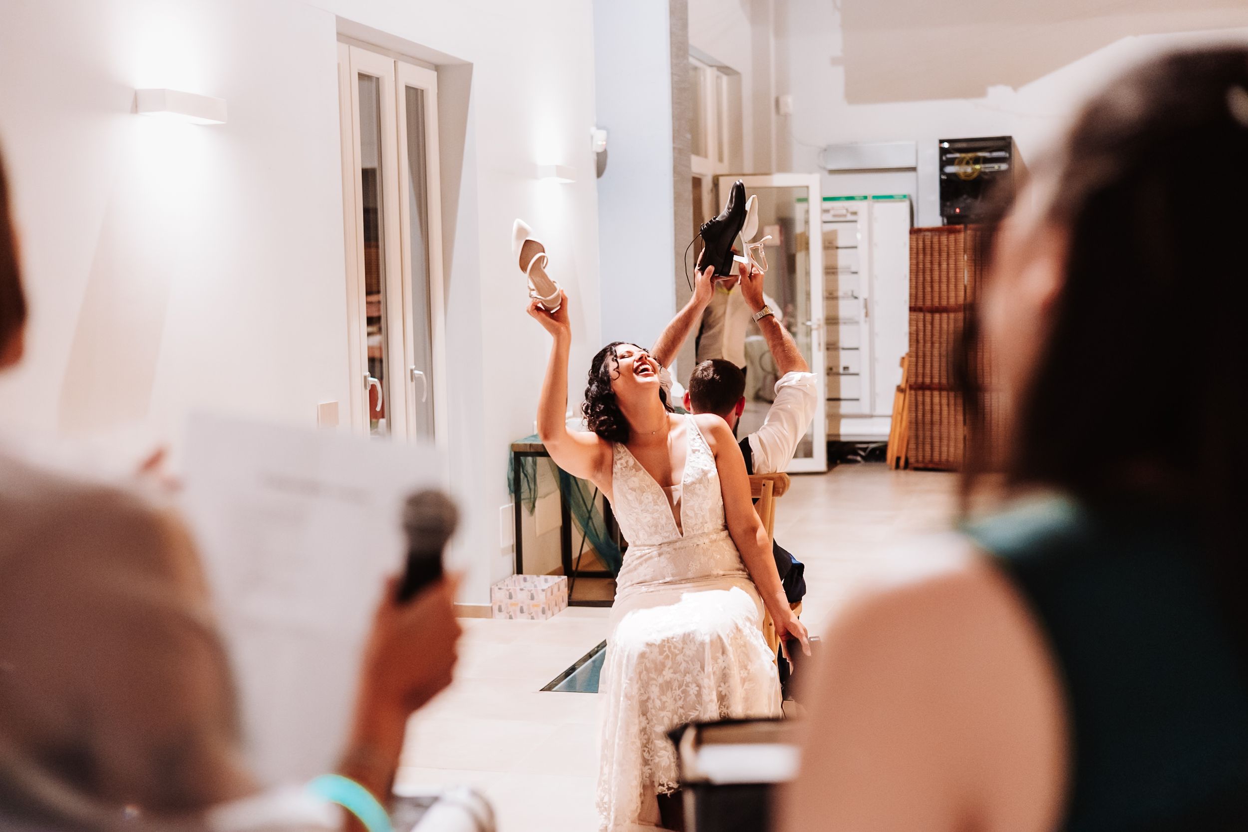 Bride in white dress joyfully raises her shoe during wedding reception celebration indoors.