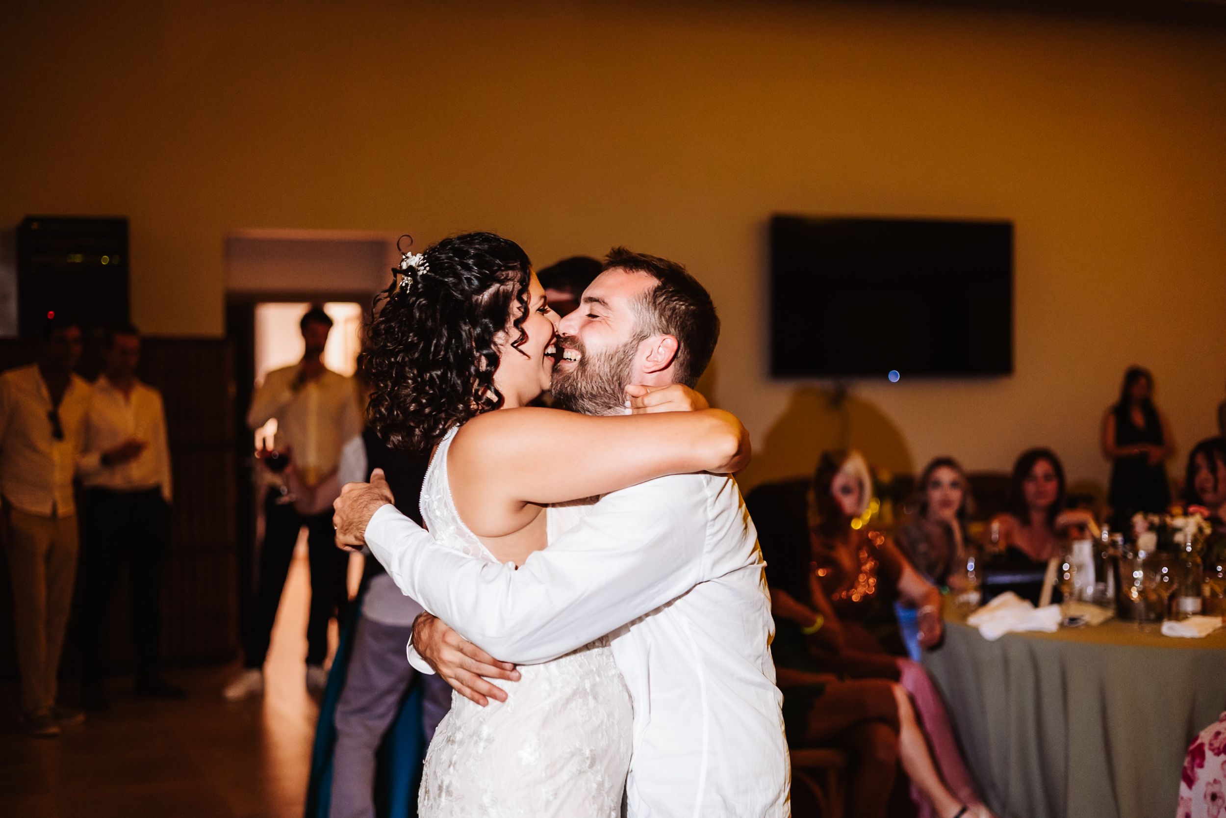 Bride and groom share romantic first dance kiss in warmly lit wedding reception hall with guests seated nearby.