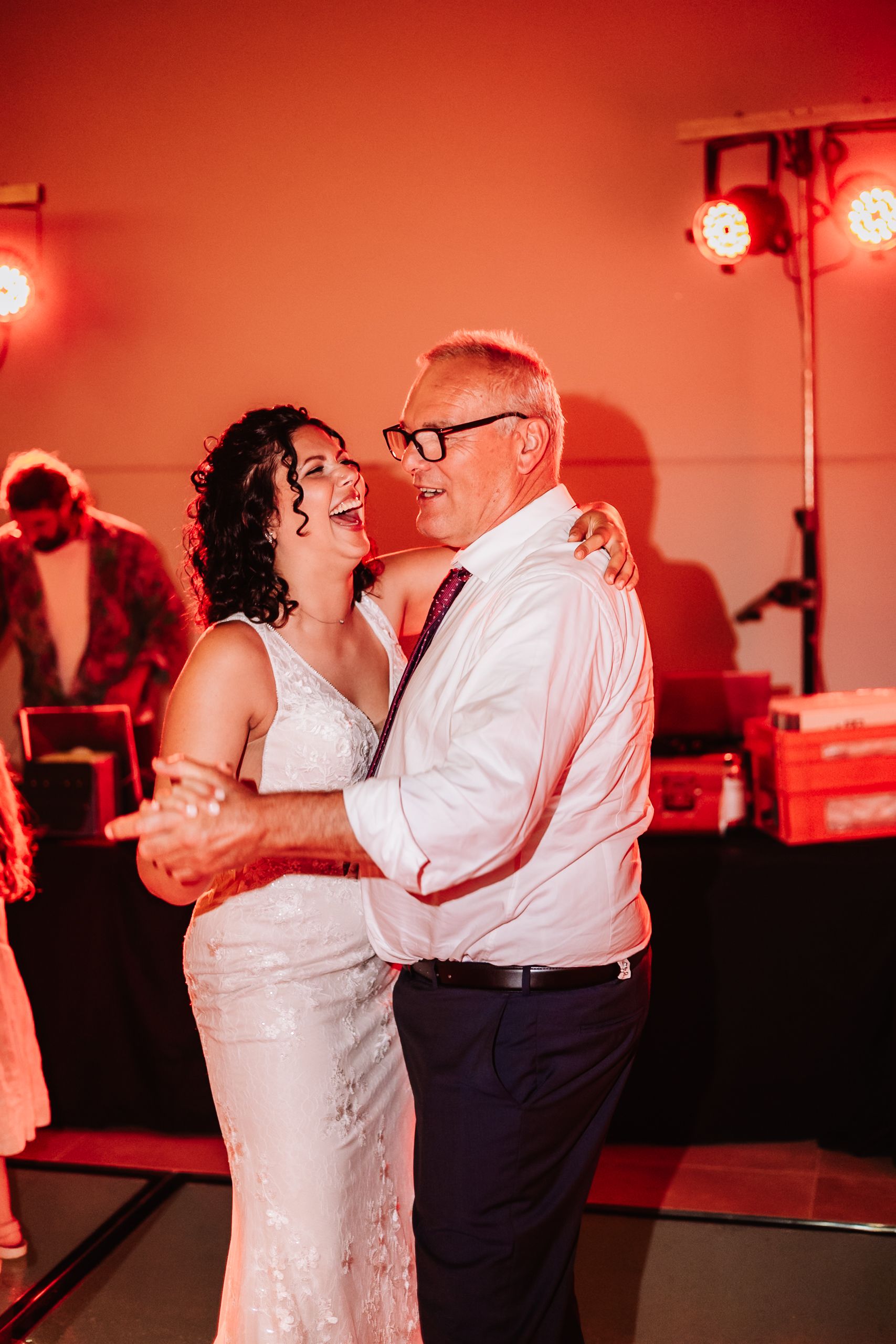 Bride in white gown dancing joyfully with older man in white shirt at wedding reception with warm red lighting.