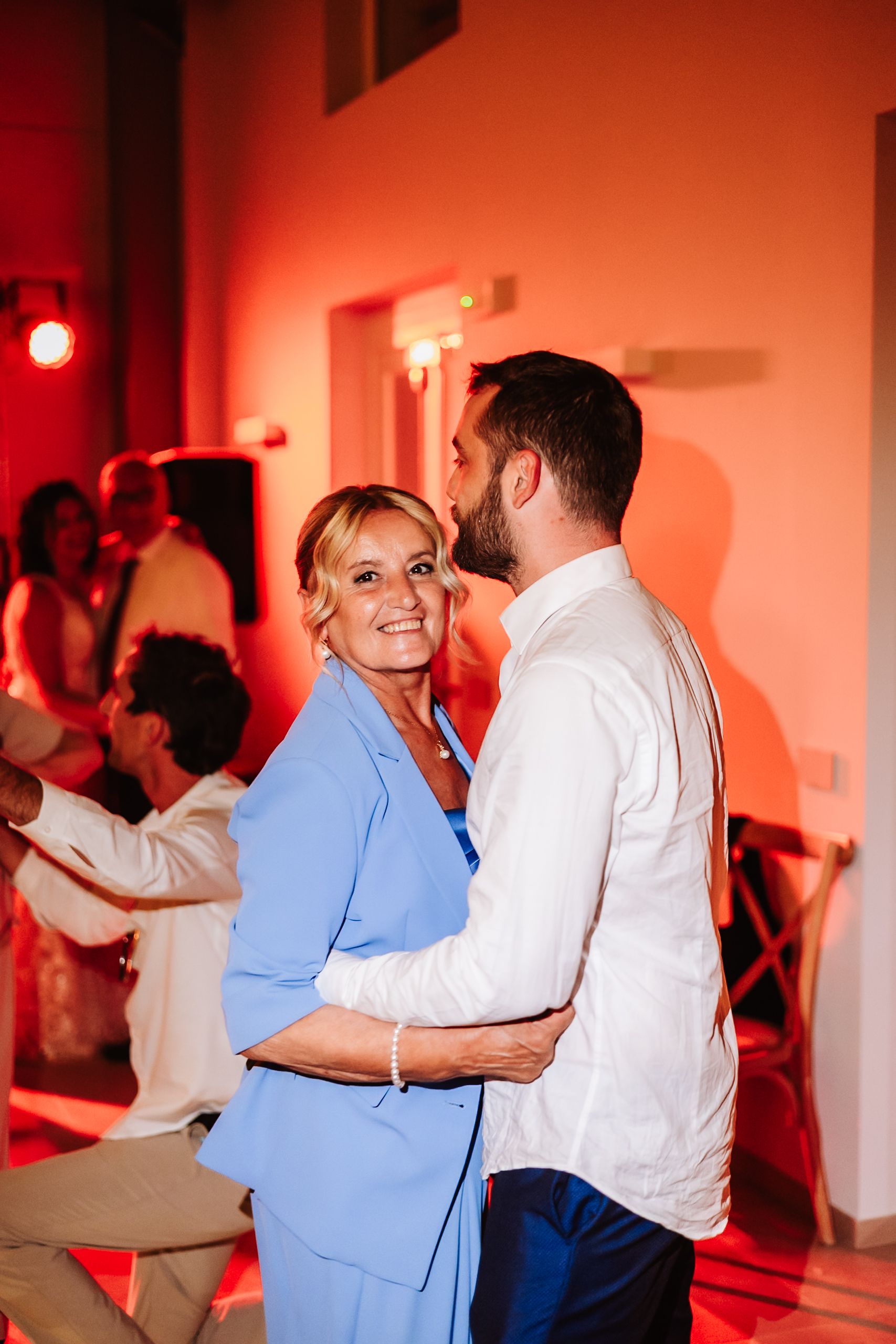 A woman in a blue suit dances with a man in a white shirt at a warmly lit celebration.
