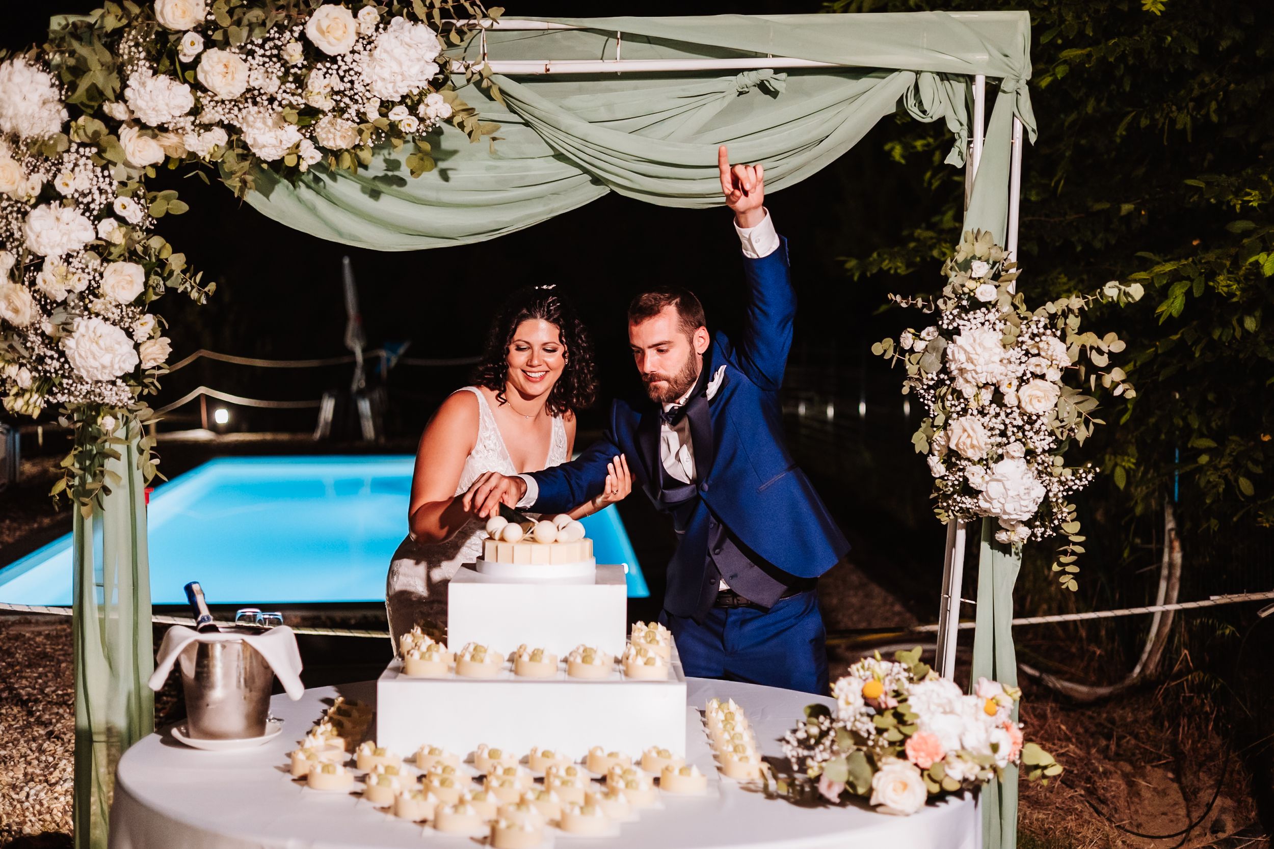 Bride and groom cutting wedding cake outdoors at night under floral arch near pool.