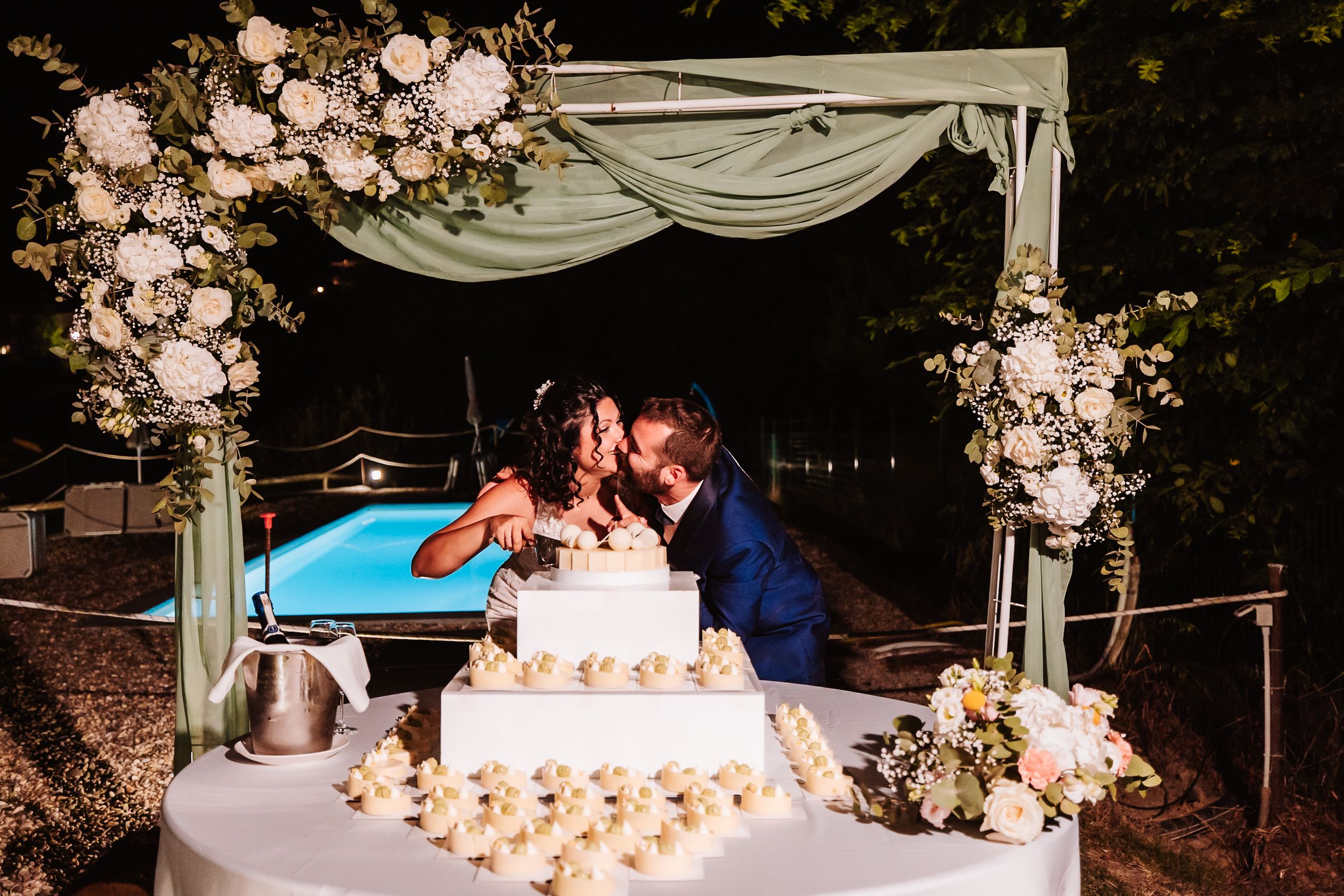 Bride and groom kissing behind a white tiered wedding cake under a floral arch with white roses at an outdoor night reception.