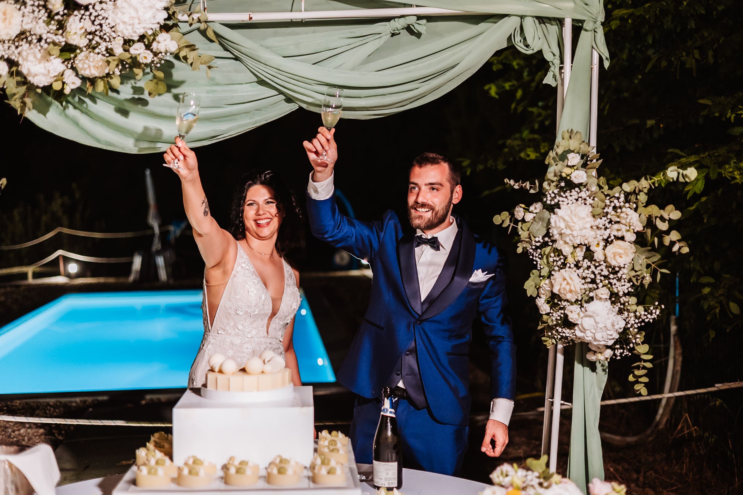 Bride and groom celebrate cake cutting under floral arch by pool at elegant outdoor wedding reception.