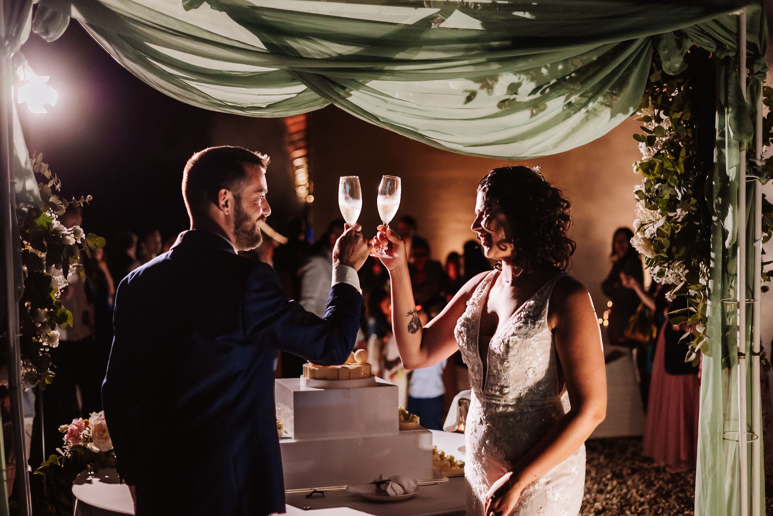 Bride and groom toasting with champagne glasses at elegant outdoor wedding reception under green draped canopy.