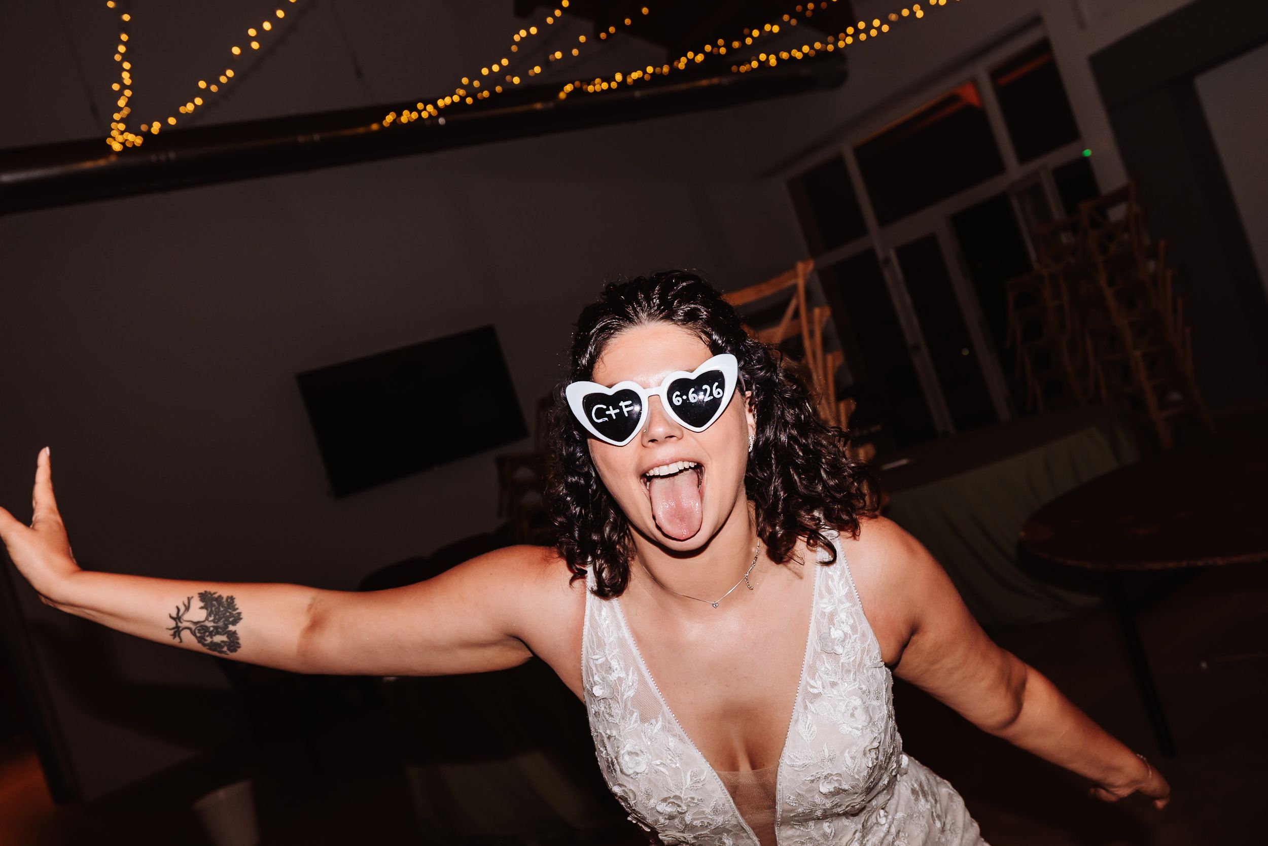 Bride in white lace dress wearing heart-shaped novelty sunglasses sticking tongue out at wedding reception.