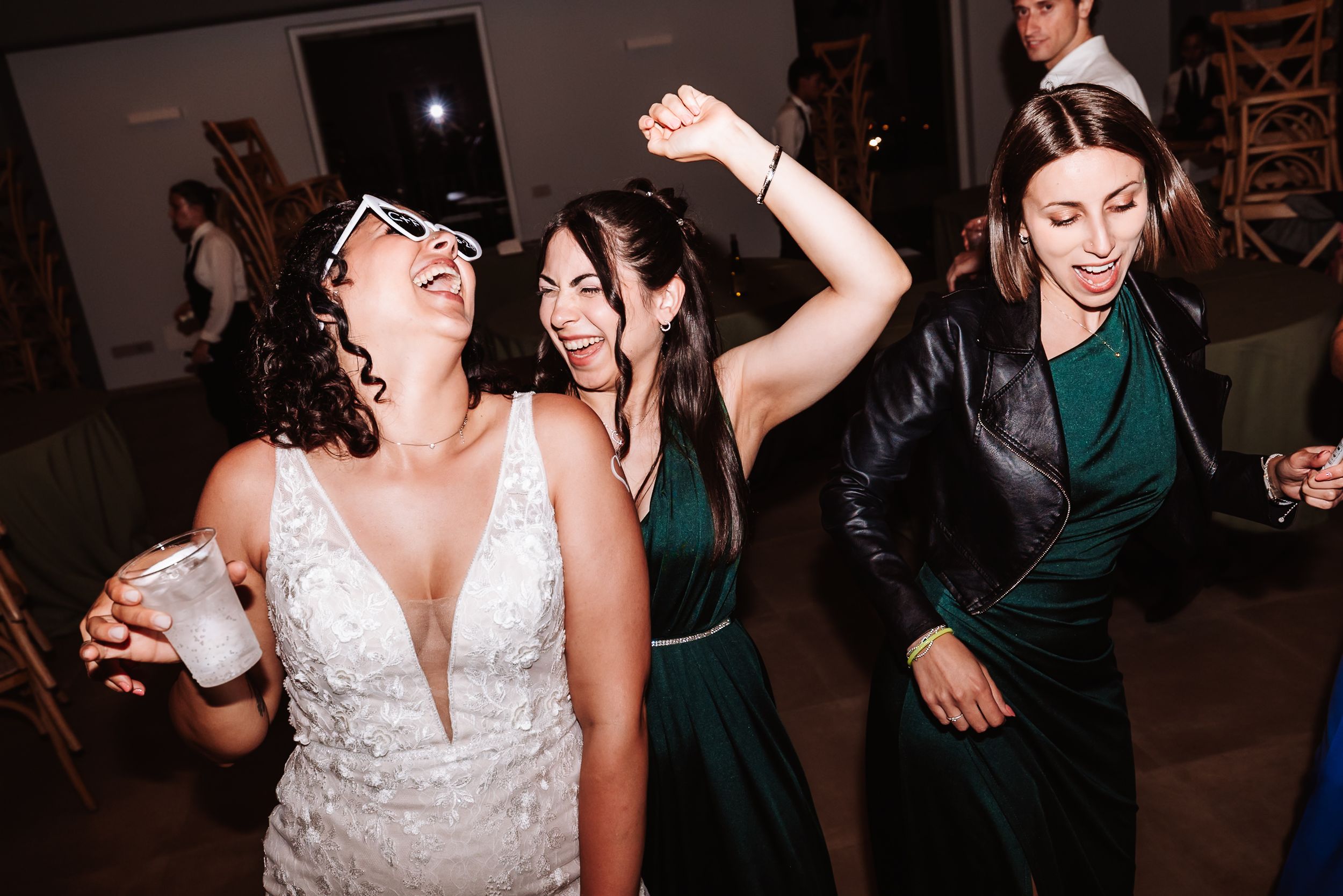 Bride and bridesmaids dancing joyfully at wedding reception, wearing white lace gown and green dresses.