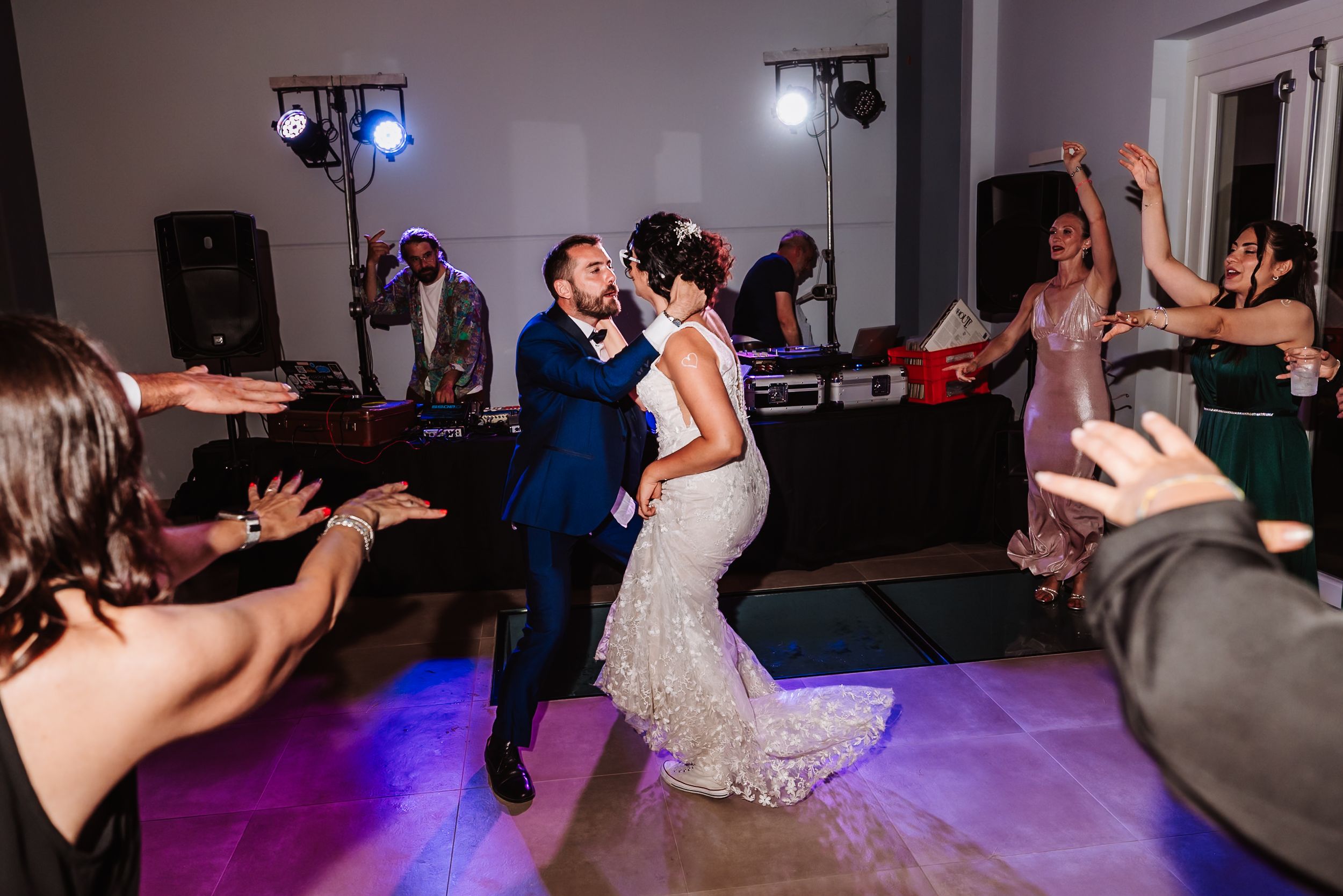 Bride and groom share first dance surrounded by cheering guests at elegant wedding reception with colorful stage lighting.