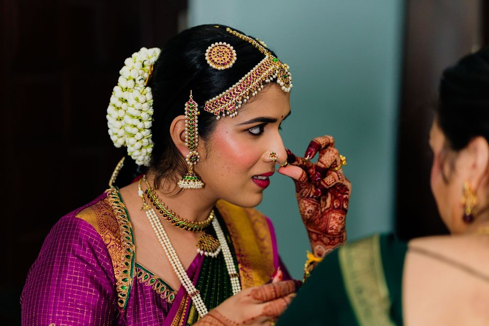Indian bride in purple silk saree with gold jewelry and henna hands being assisted during traditional wedding ceremony.