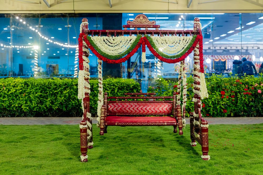 Ornate Indian wedding swing decorated with red and white flower garlands on green lawn.