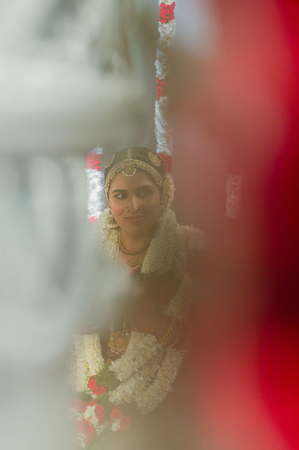 Indian bride in traditional bridal jewelry and floral garlands seen through a soft focus frame.