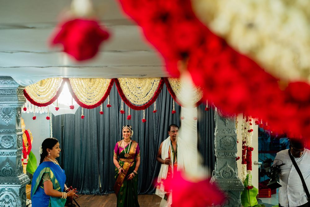 South Indian bride in colorful silk saree stands with groom at decorated wedding mandap with red floral backdrop.