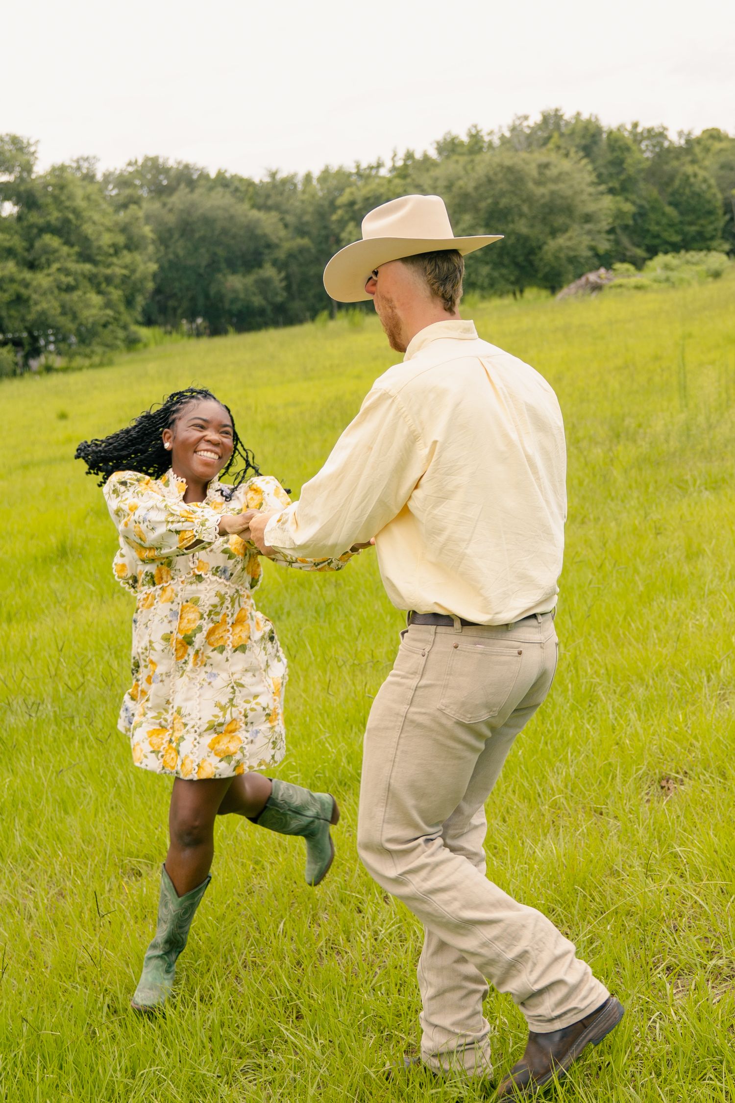 A joyful couple dances in a lush green meadow, woman in floral dress and man in cowboy hat holding hands.