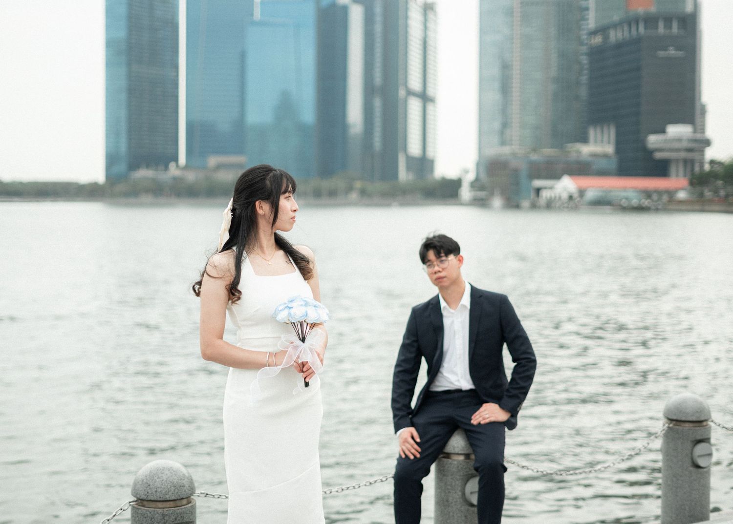 A couple in wedding attire share a playful moment together on a lawn with white buildings in the background.