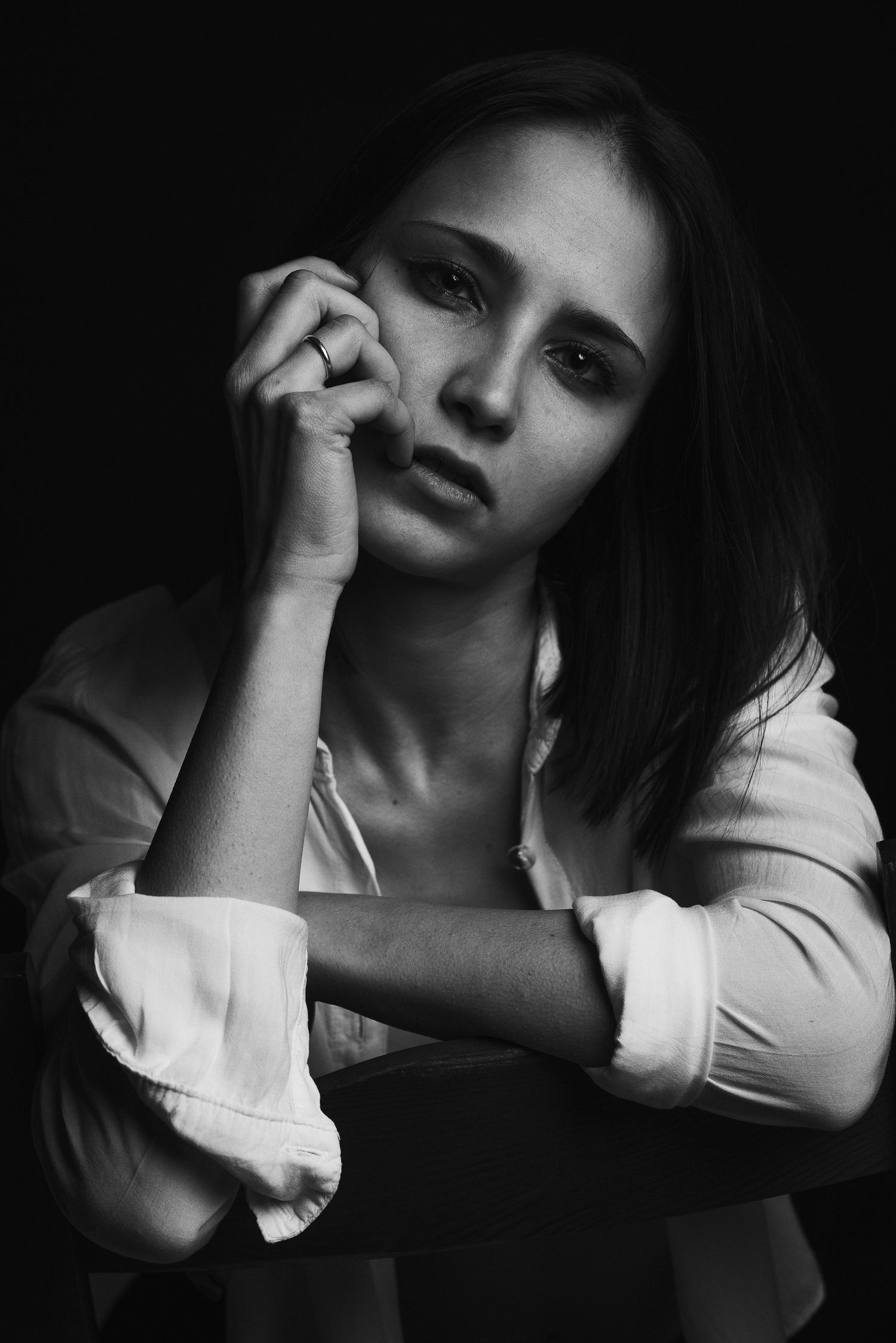 Primo piano espressivo in bianco e nero di una donna con camicia bianca, appoggiata allo schienale di una sedia con la mano vicino al mento