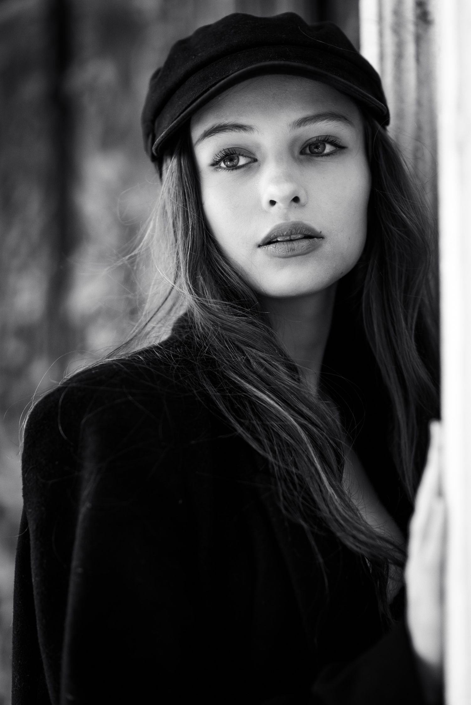 Primo piano in bianco e nero di una ragazza con cappello stile marinaio e cappotto scuro, appoggiata a una parete mentre guarda di lato.