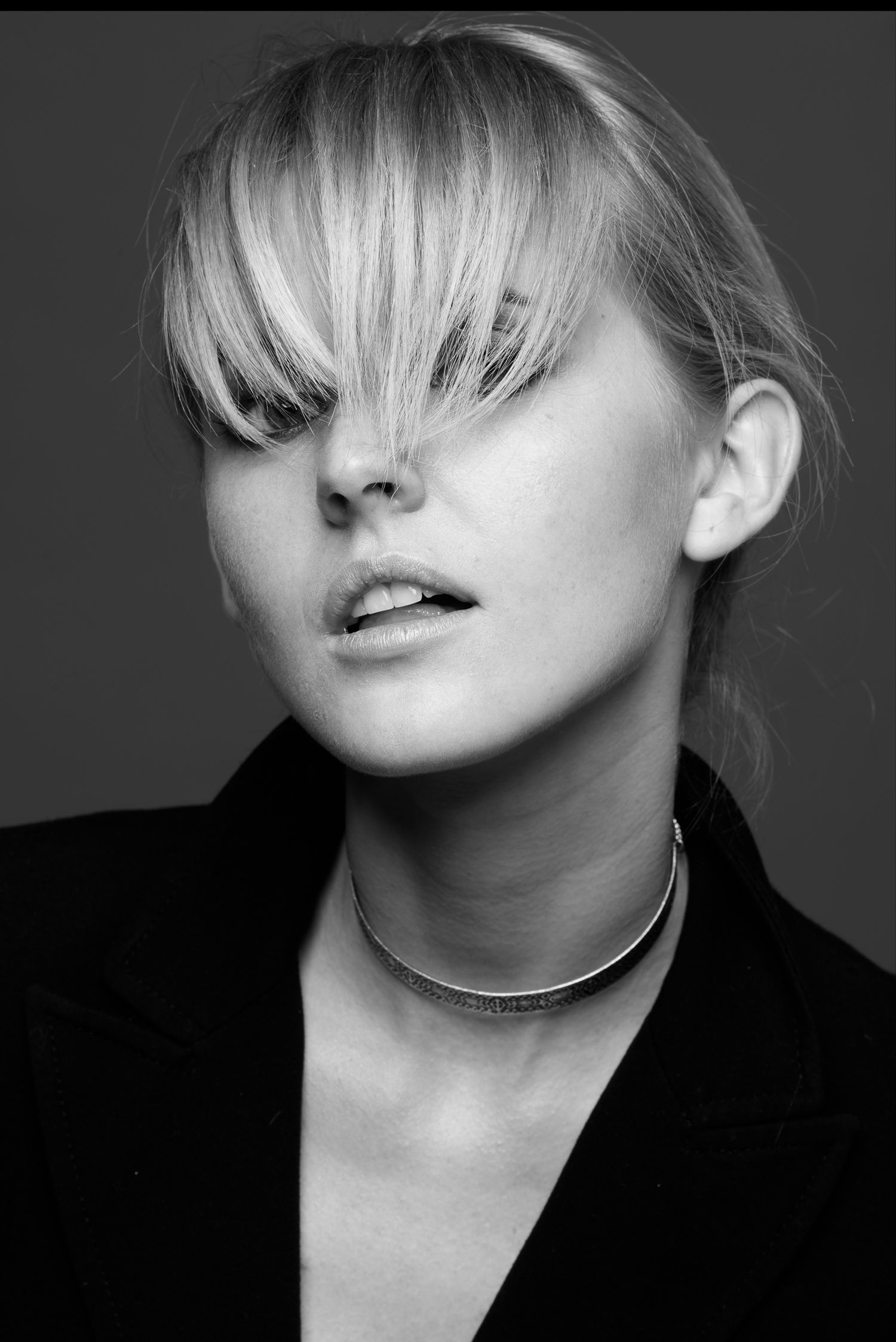Primo piano glamour in bianco e nero di una modella con lunga frangia bionda che le copre parzialmente gli occhi, collana choker sottile e g